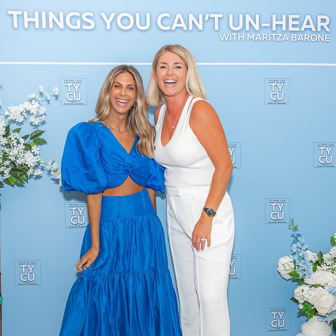 Two women smiling and posing together in front of a blue backdrop with floral decorations. One woman is wearing a blue dress with puffy sleeves, and the other is wearing a white sleeveless outfit. The backdrop has the text 'THINGS YOU CAN'T UN-HEAR WITH MARITZA BARONE' and the logo 'TYCU'.