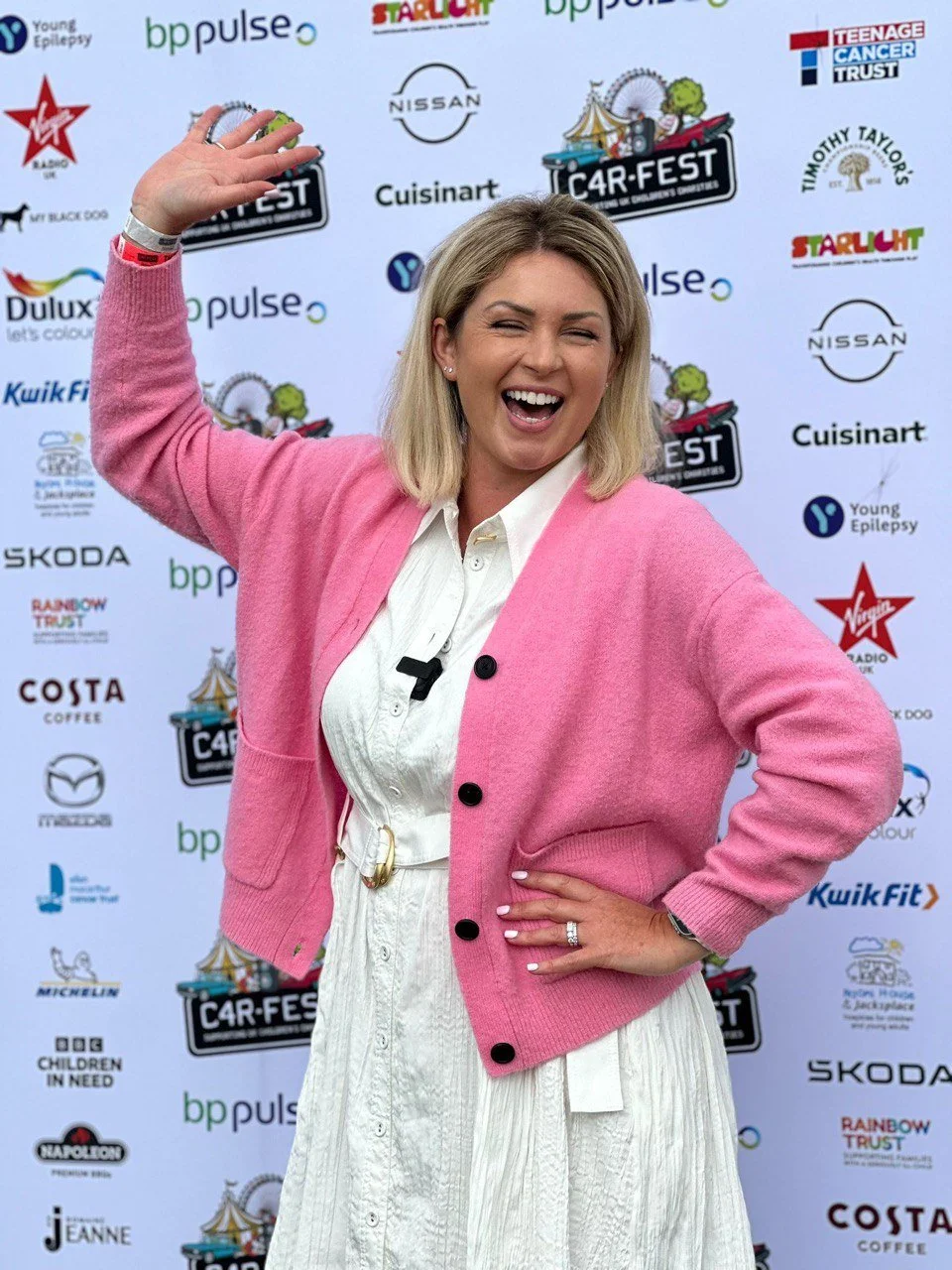 Kimberly Gallagher wearing a pink cardigan and a white dress, smiling and waving in front of a backdrop featuring various sponsor logos and event names.