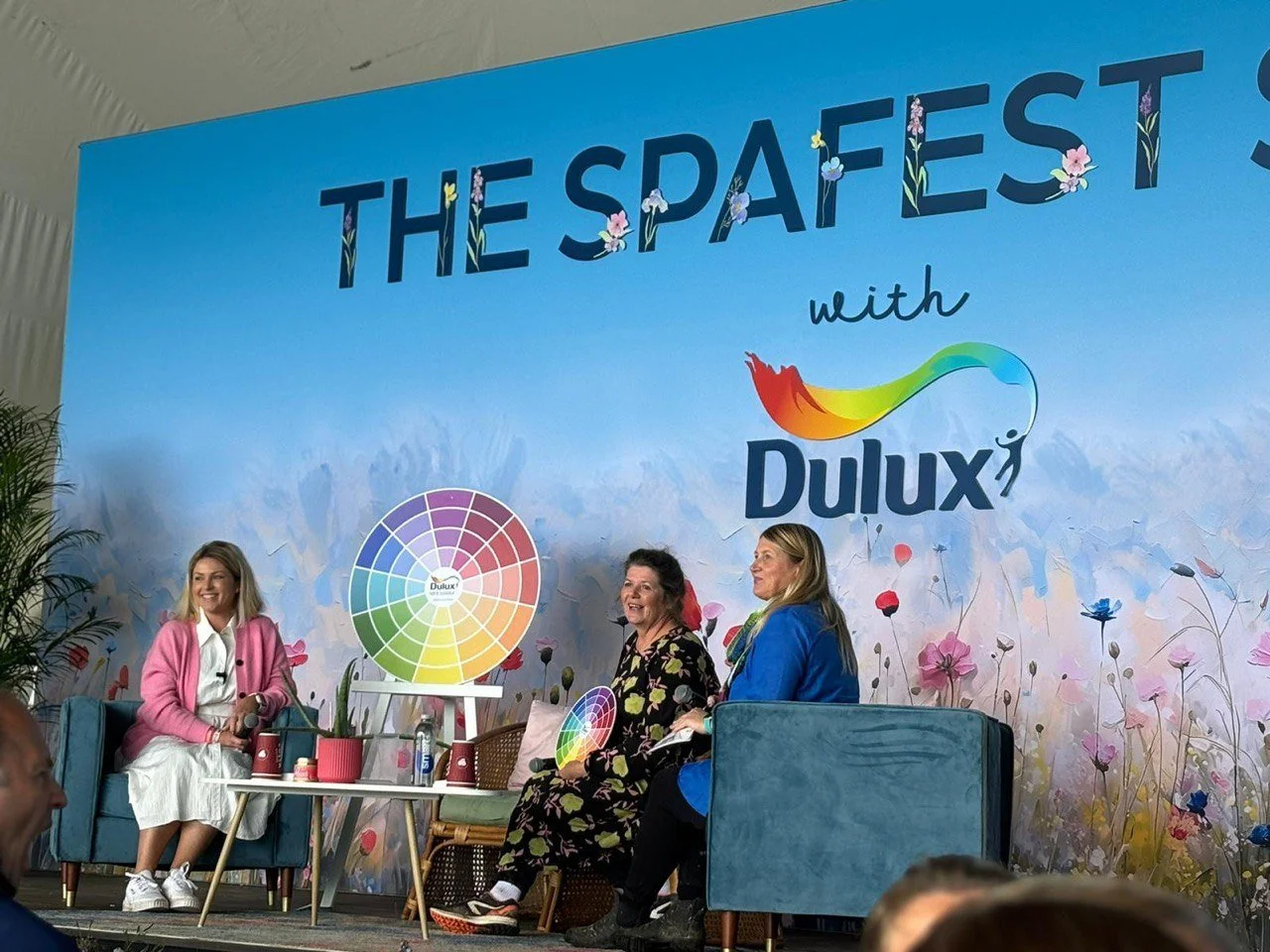 Kimberly Gallagher  with two other women sitting on chairs on stage at the SPAFEST event, with a colourful Dulux logo and a colour wheel in the background, along with floral decorations.