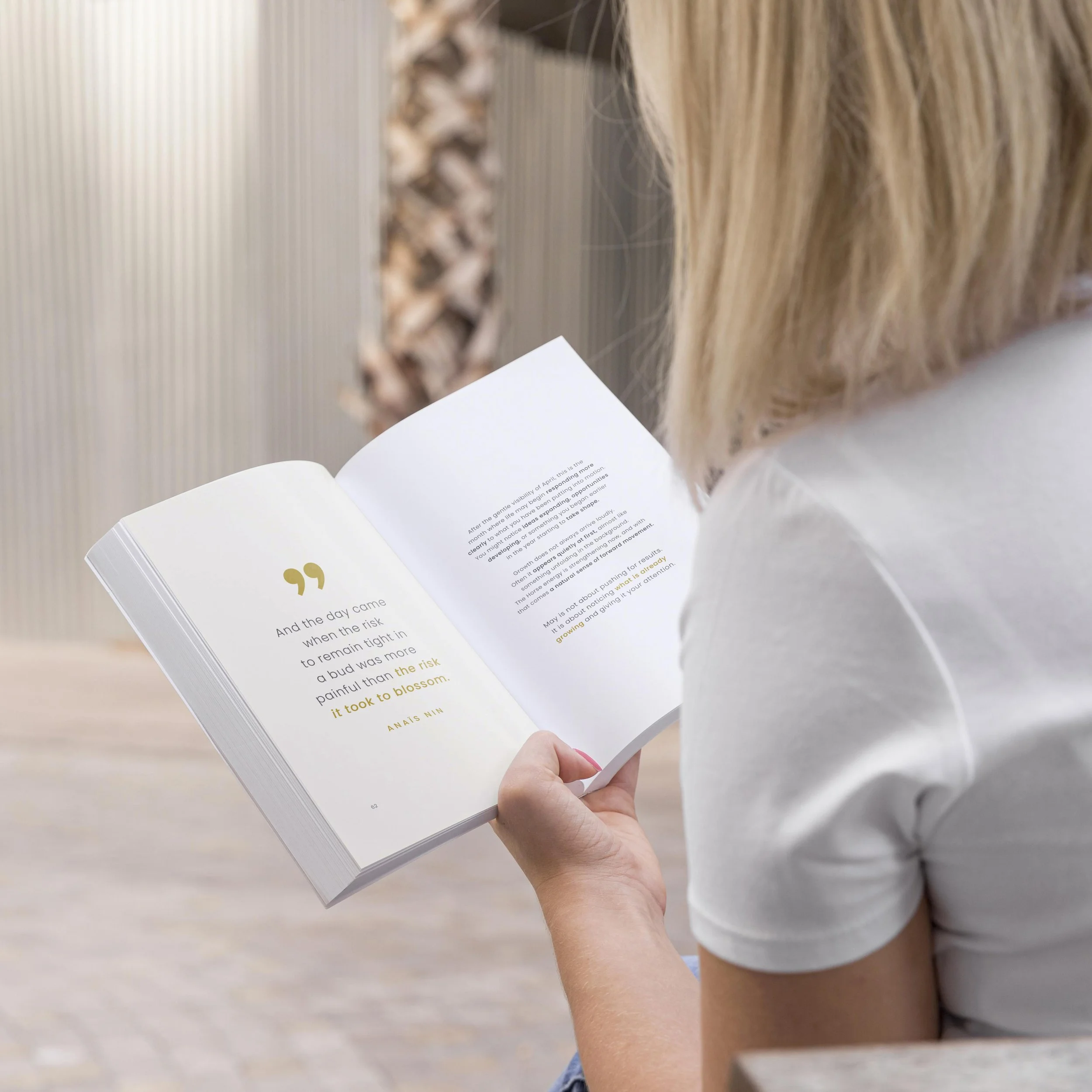 A person holding an open book with typed text and yellow quotation marks on a white page, with long blonde hair and a white shirt.
