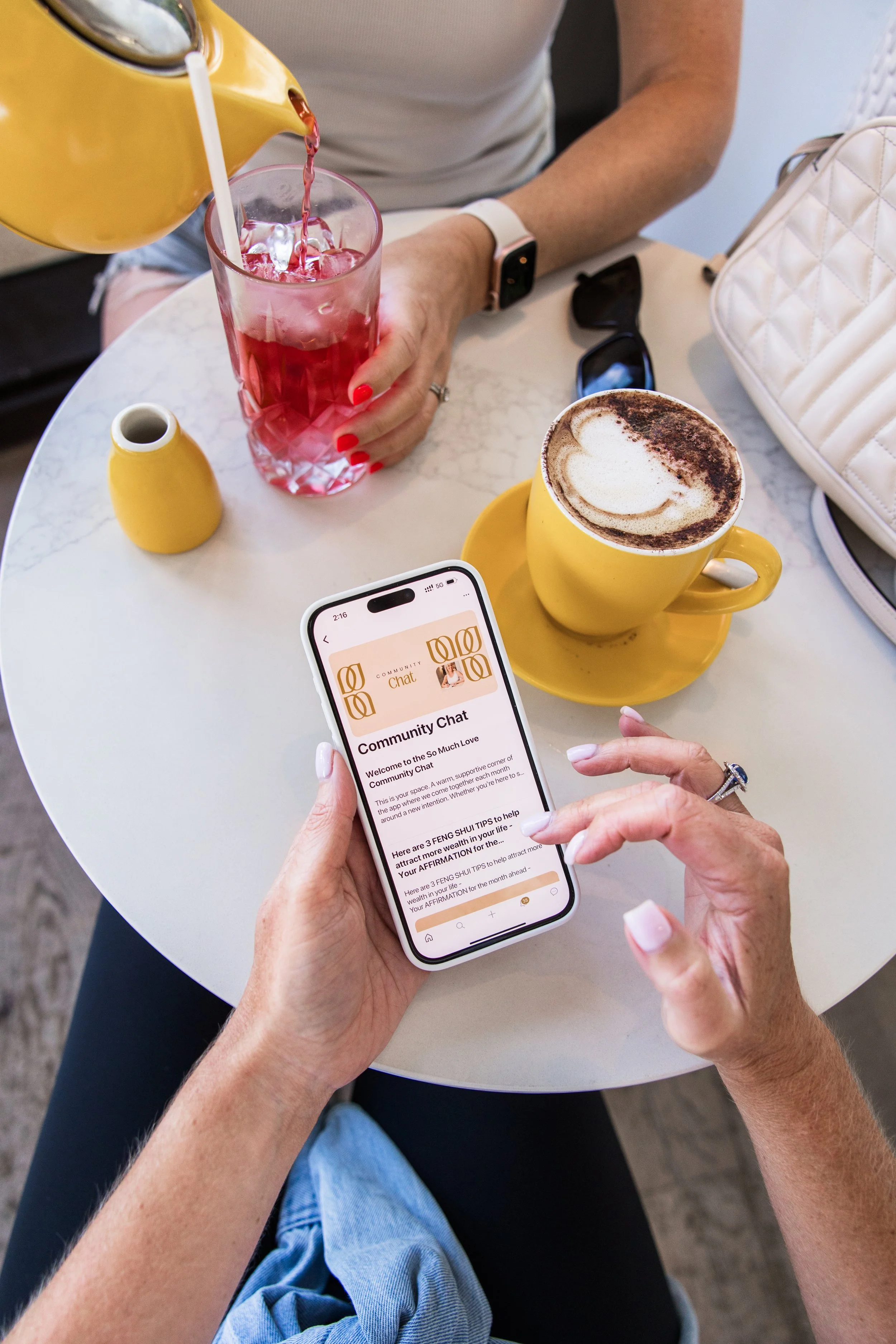 Top view of a person holding a smartphone displaying a chat app, sitting at a round white table with a yellow coffee mug, a glass of pink drink, a small yellow vase, a pair of sunglasses, and a white quilted bag.