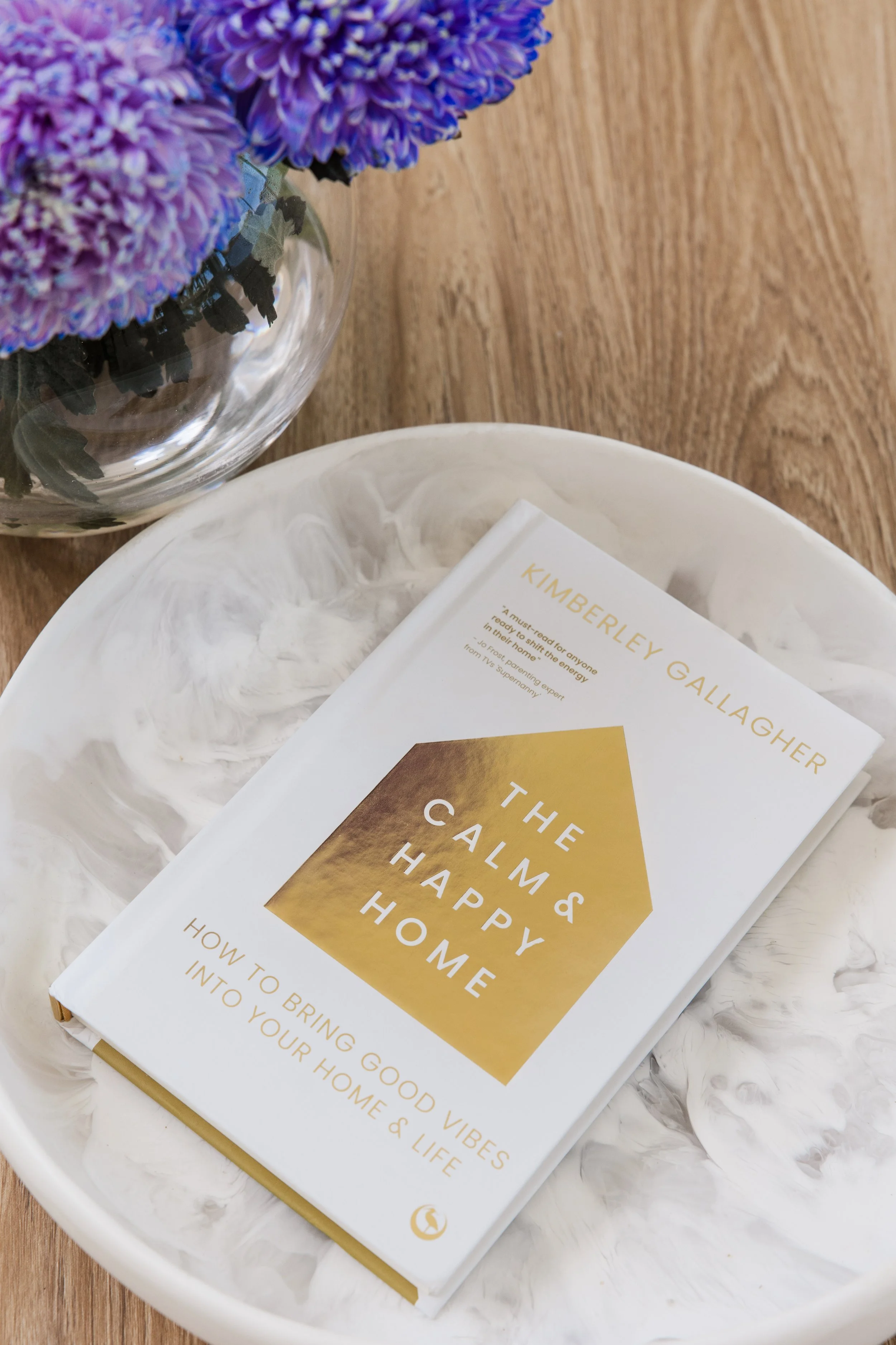 A white marble tray holding a book titled "The Calm & Happy Home" by Kimberley Gallagher and a clear glass vase with purple chrysanthemums, on a wooden surface.