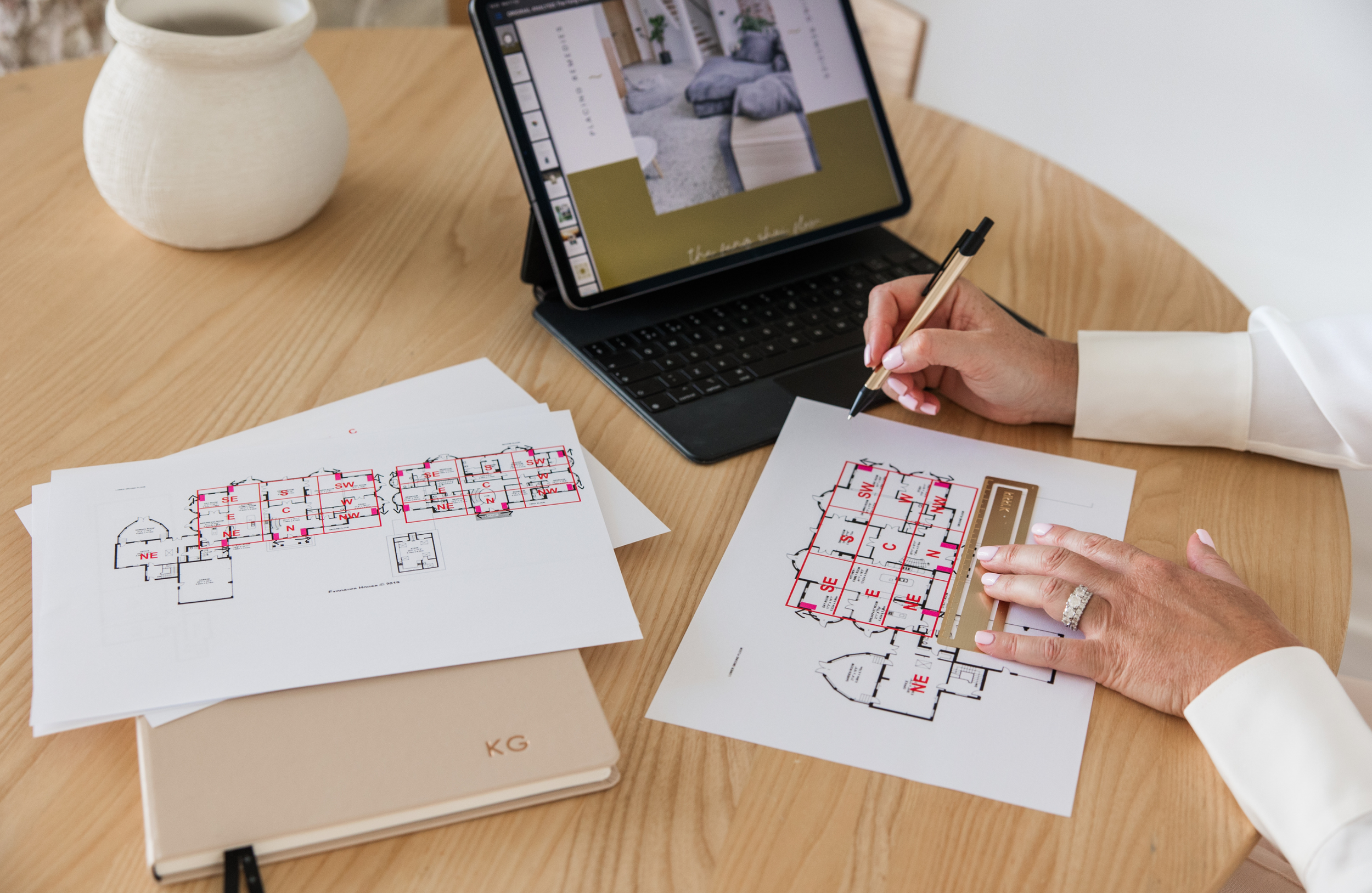Person working on architectural floor plans at a wooden table with a tablet displaying a room interior and various papers with building layouts.