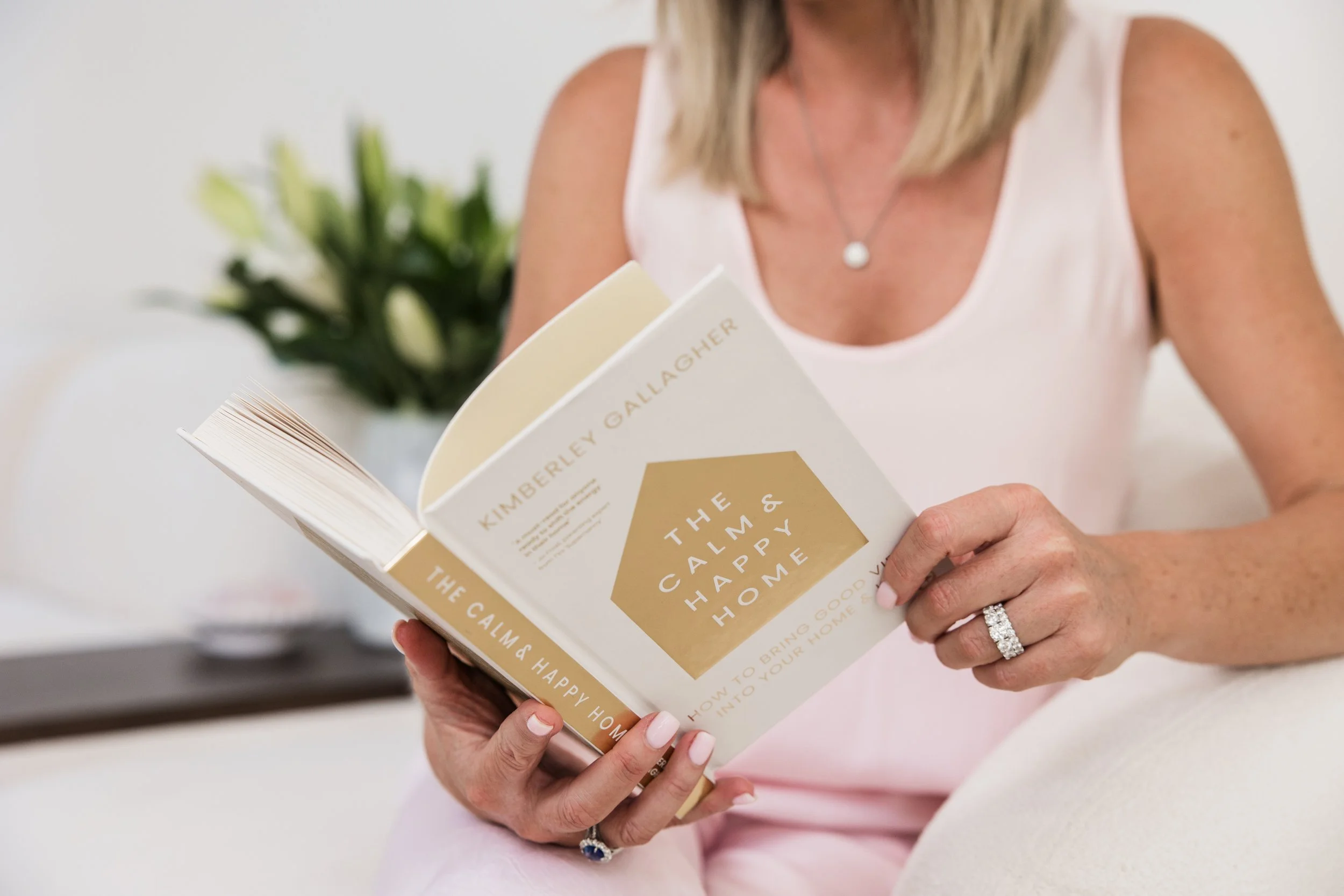 A woman with blonde hair wearing a white sleeveless top and a silver necklace is reading a book titled 'The Calm, Happy Home.' She has a wedding ring on her left hand.