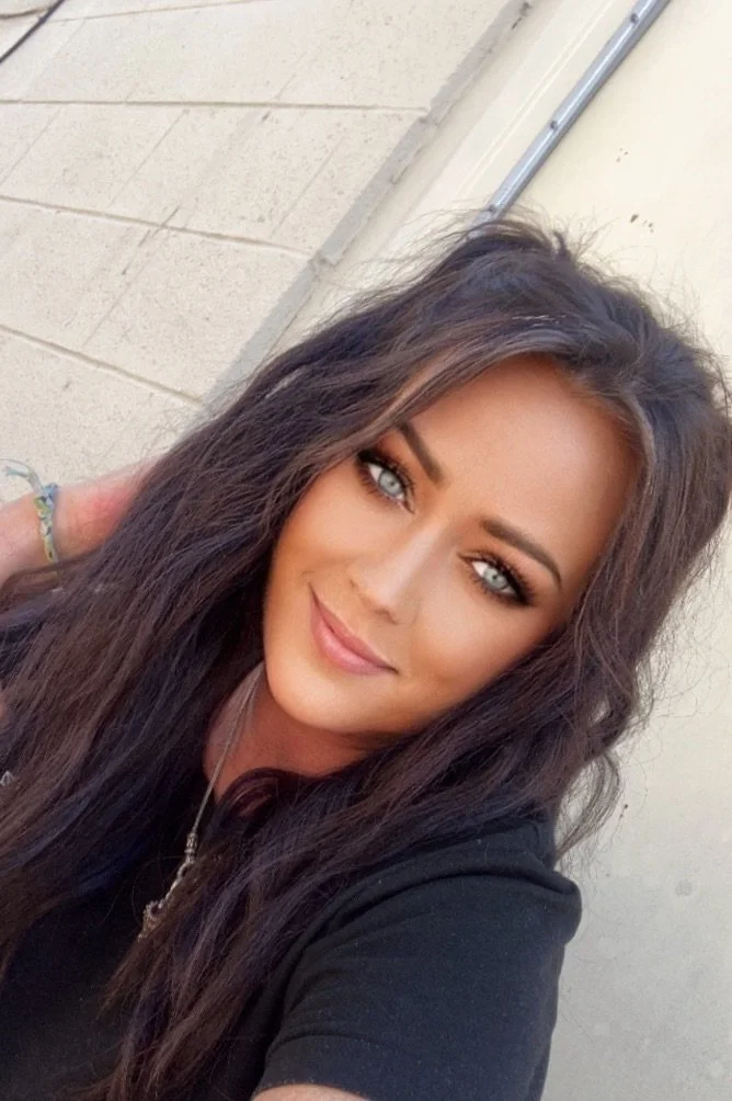 A woman with long dark hair, blue eyes, and makeup, smiling in front of a beige wall.