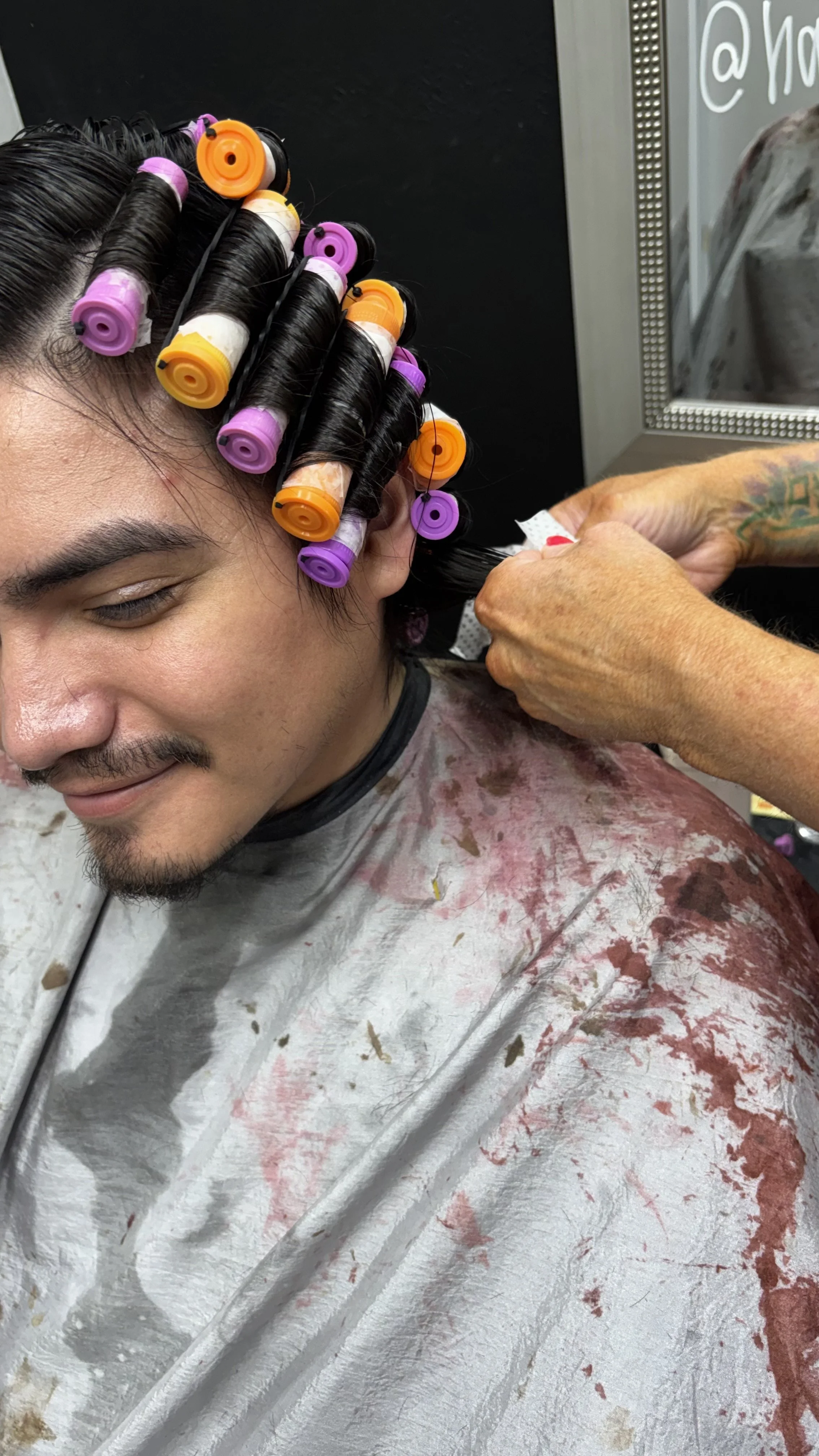A person getting their hair colored with hair rollers in a salon, smiling with eyes closed, and wearing a smock stained with hair dye.