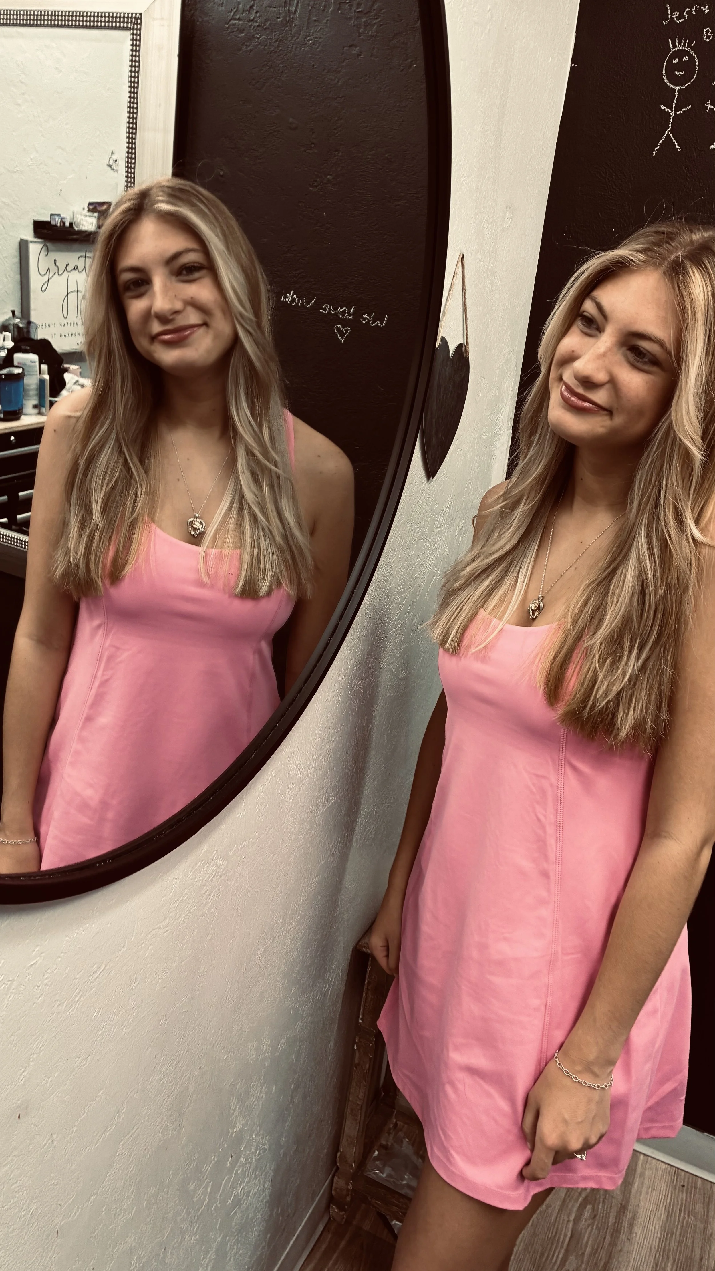 A young woman with long, wavy blonde hair wearing a pink dress and jewelry, standing in front of a mirror inside a room. The room has a black chalkboard wall with white writing and drawings, and a white wall with some writing and decorations. Her reflection is visible in the mirror.