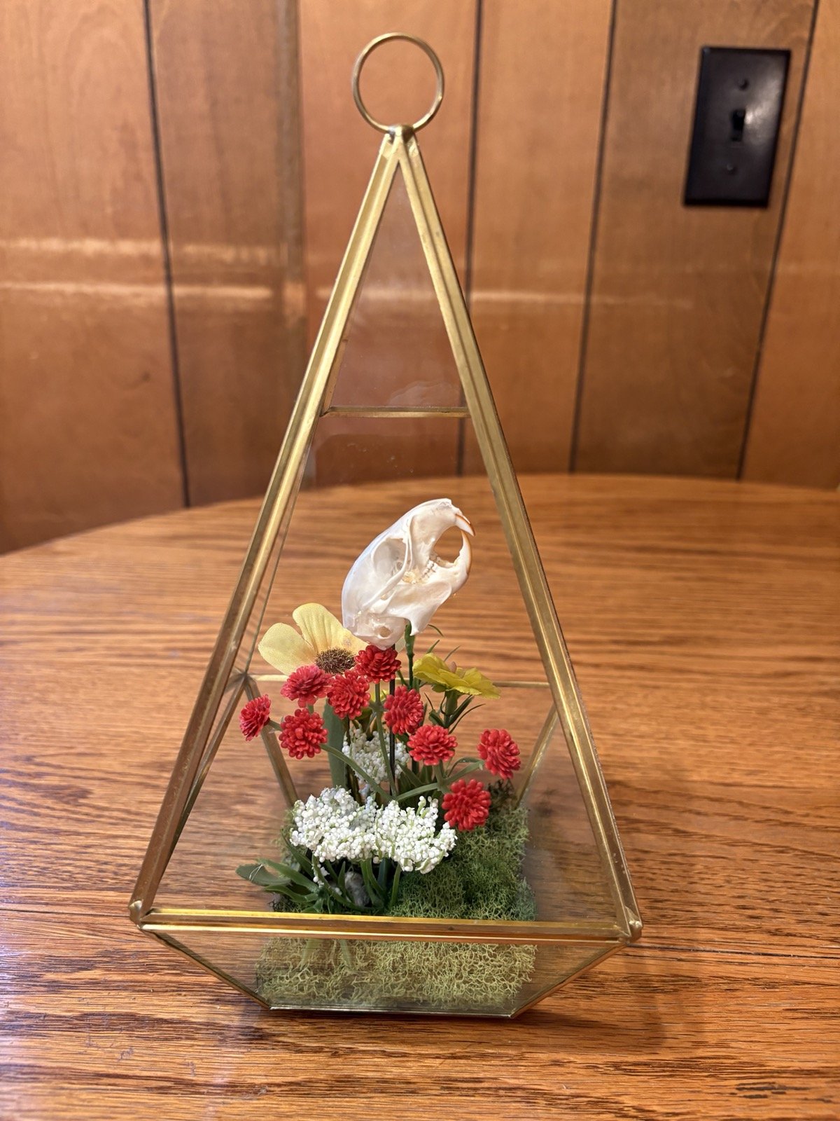 A terrarium with a gold triangular frame containing a small animal skull and colorful flowers on a wooden table.