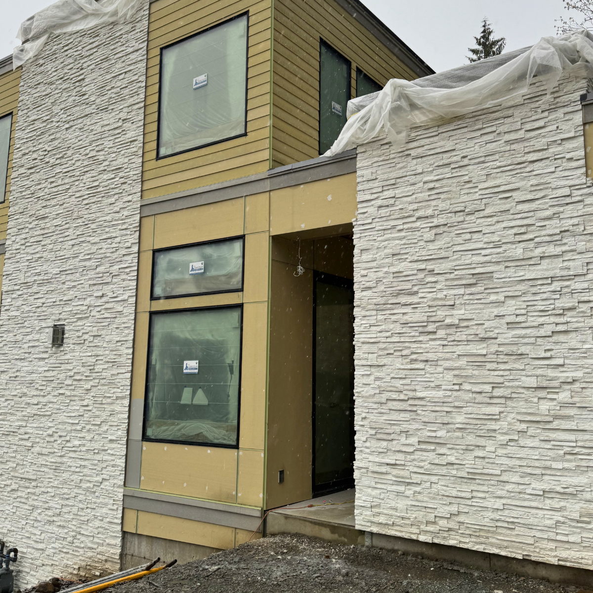 Exterior view of a modern, multi-story building under construction, featuring textured white stone wall panels and yellow siding, with large window openings covered with protective plastic, and construction materials on the ground.