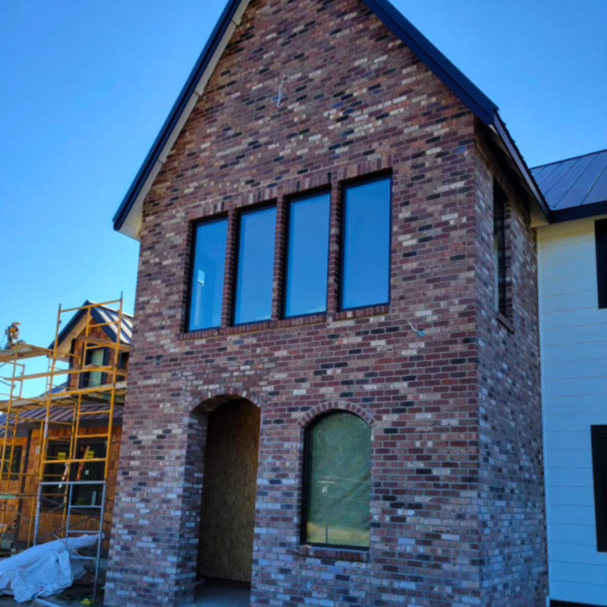 A three-story brick house under construction, with three large windows on the upper floor and an arched doorway on the ground floor, with other houses and construction scaffolding nearby.