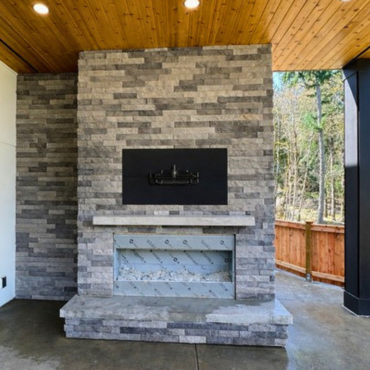 Outdoor stone fireplace under a wooden ceiling on a patio with a view of trees and a wooden fence.