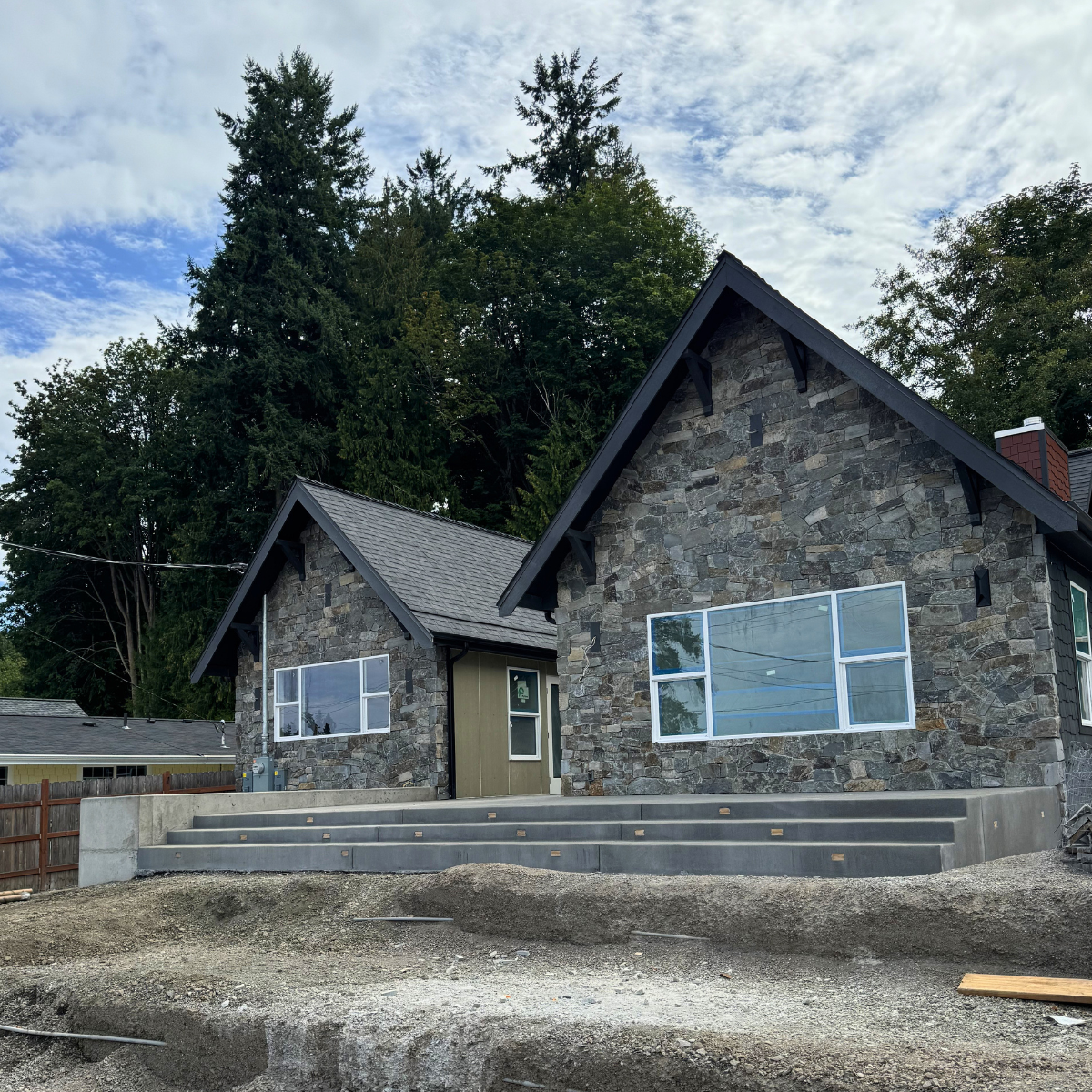 Stone house with large windows and front steps under a partly cloudy sky, surrounded by trees.