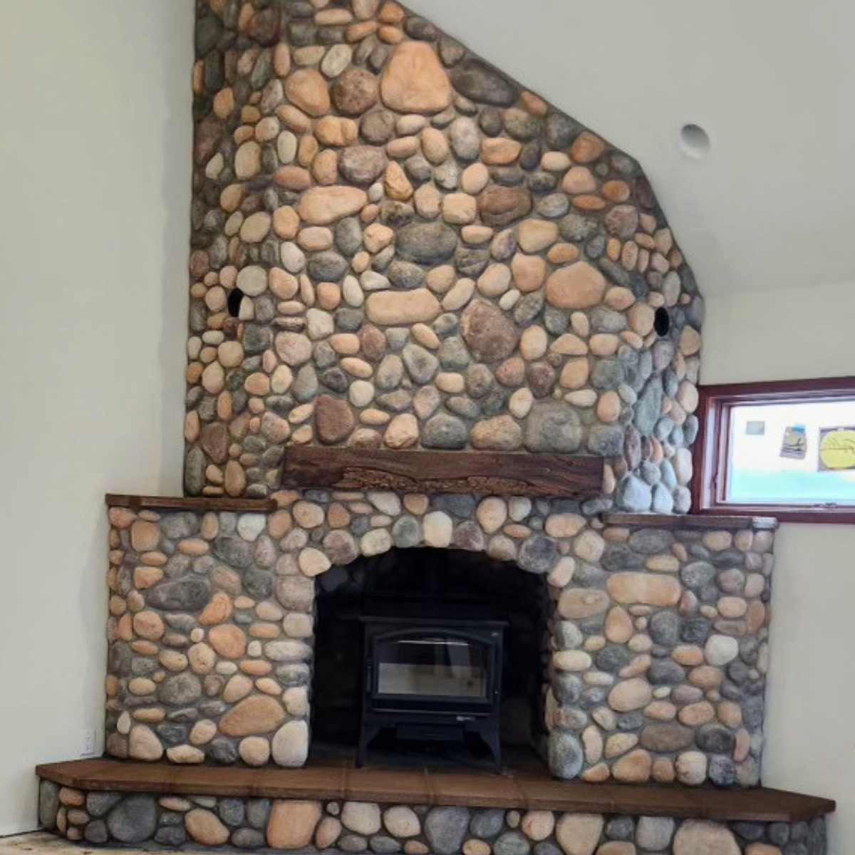 A large stone fireplace with a wood mantel and a black stove insert, surrounded by multicolored smooth stones, in a room with a small window.