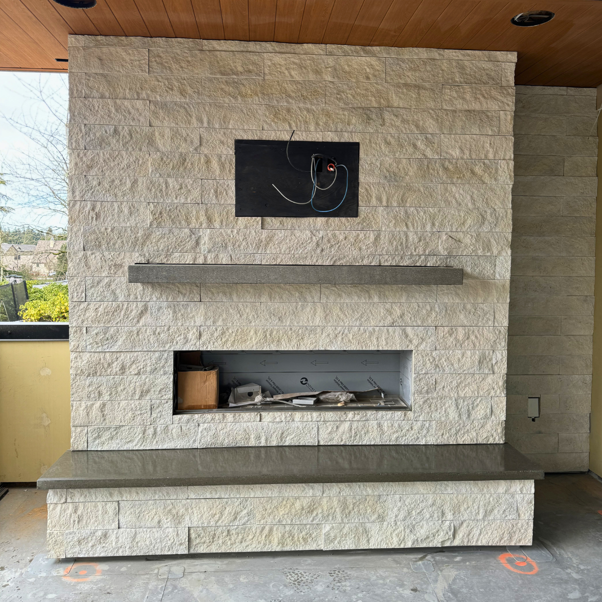 Unfinished fireplace with wiring and a black wall-mounted panel, surrounded by beige stone tiles, in a room under construction.