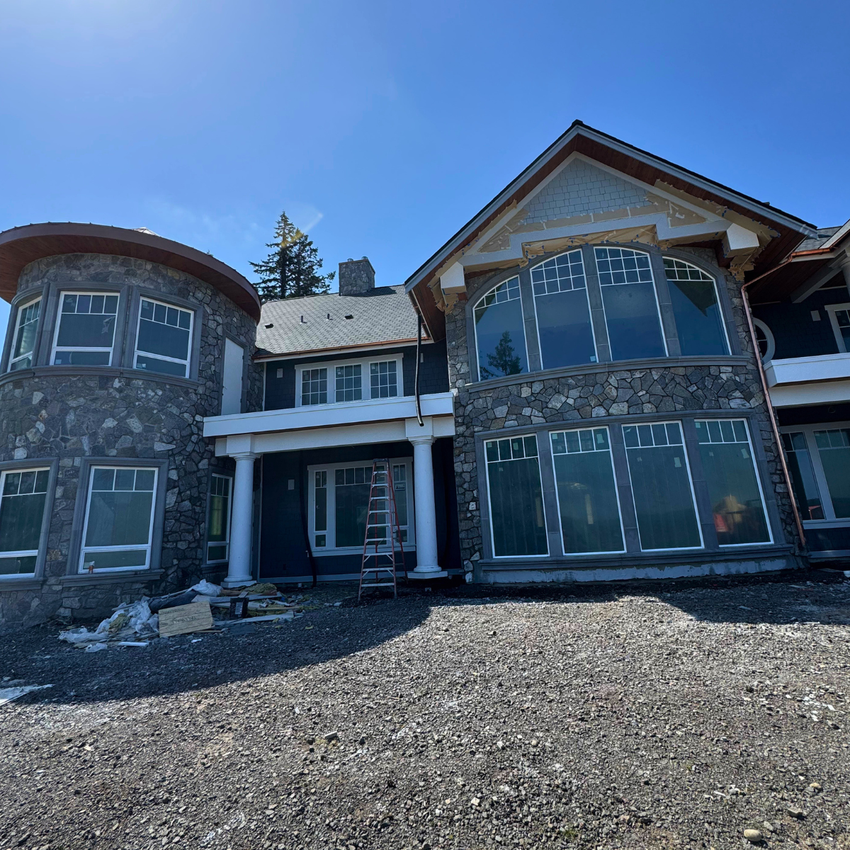 A large, multi-story house under construction with stone and siding exterior, multiple large windows, and a prominent front porch supported by white columns, with a clear blue sky overhead.
