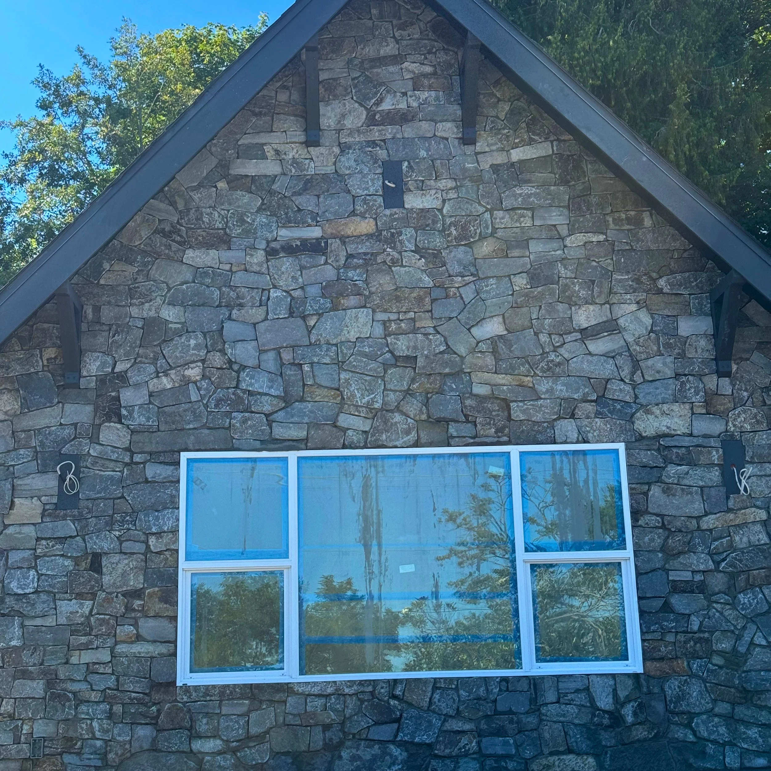 A building with a stone facade and diamond-shaped black trim on the roof peak, reflecting trees in a large window, under a clear blue sky.