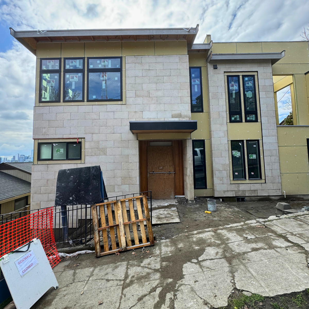 Under construction modern multi-story house with stone exterior walls, large windows, and a temporary wooden door; construction materials and signs indicating closed sidewalk in front.