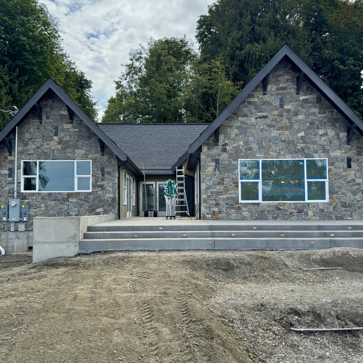 A house under construction with a stone exterior, large windows, and a newly built concrete porch with steps. Construction tools and ladder are visible on the porch. Dirt surrounds the house, and trees are in the background under a cloudy sky.