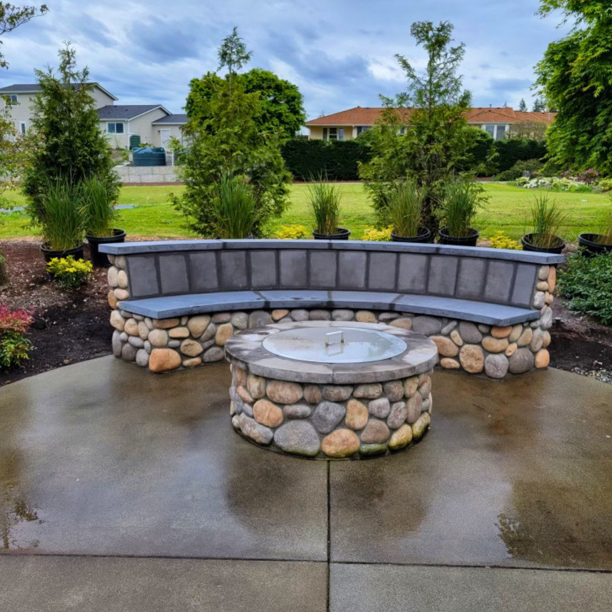 A curved stone bench and a round stone fire pit with a water feature in a backyard garden, with potted plants and trees in the background under a cloudy sky.