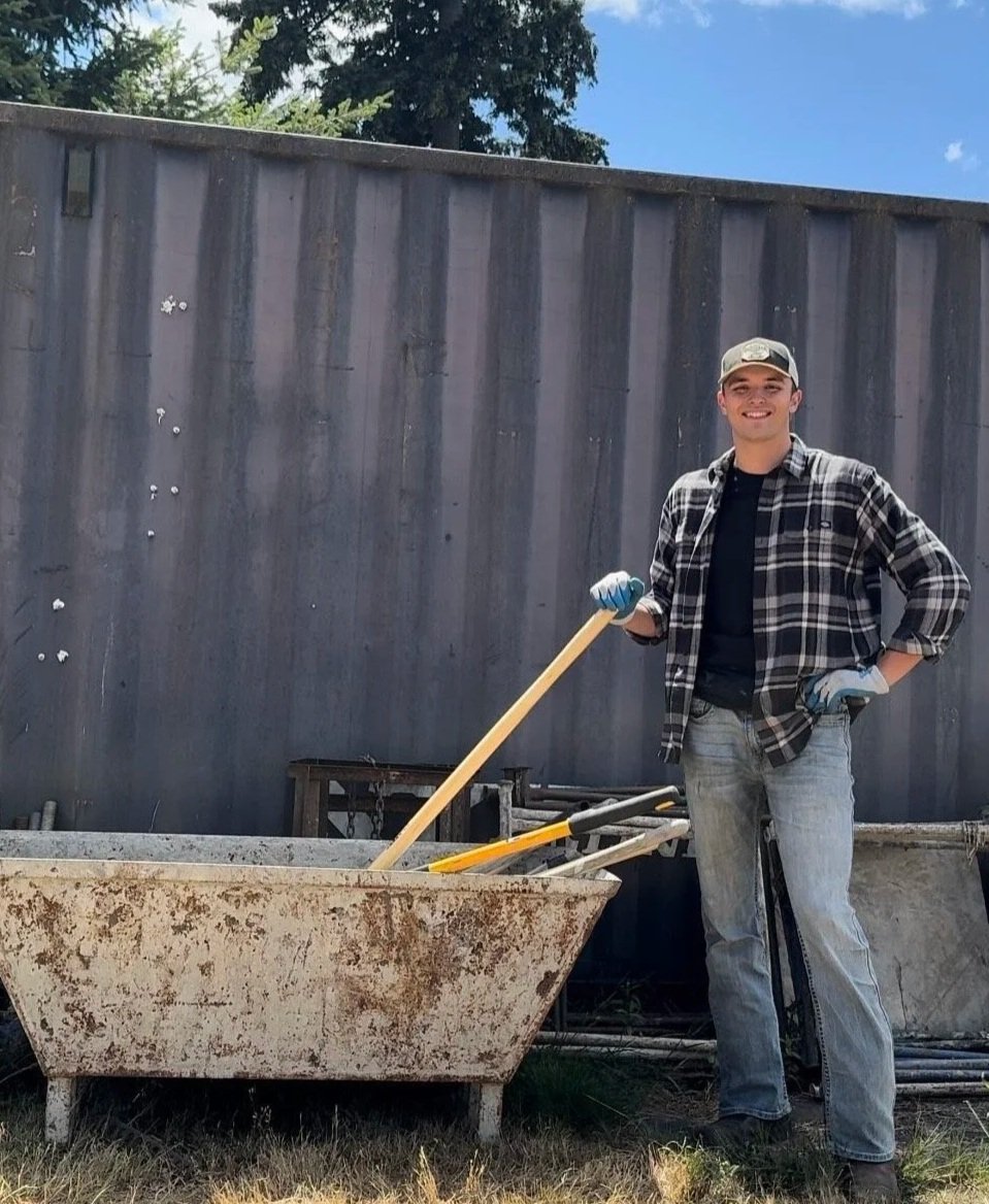 A young man in a plaid shirt, jeans, and a baseball cap stands outdoors next to a rusty bathtub filled with tools, holding a shovel, with a dark wooden fence and blue sky in the background.