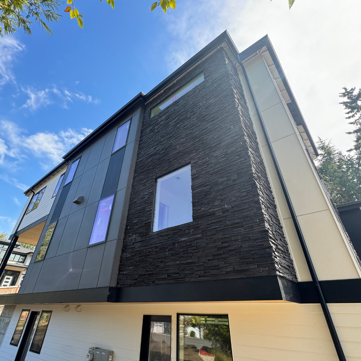 Modern multi-story house with a combination of gray, white, and black exterior siding, large windows, and a black gutter, situated under a blue sky with some clouds.