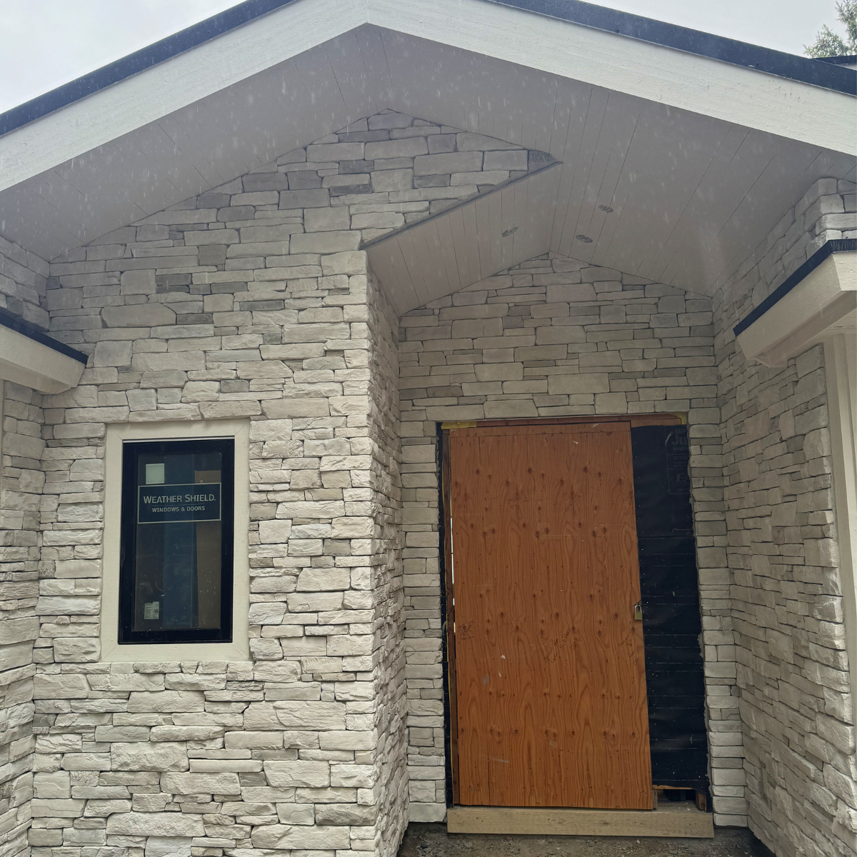 Front entrance of a house under construction with stone siding, a small window to the left, and a wooden door with plywood covering the entrance.