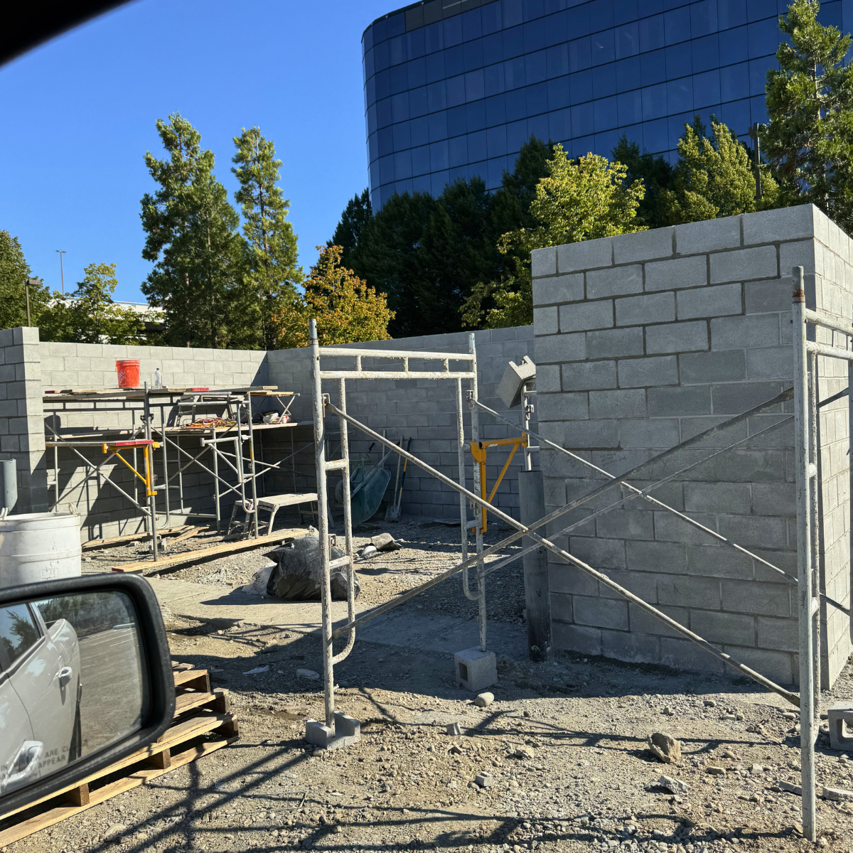Construction site with concrete block walls, scaffolding, and construction tools, with a modern glass building and trees in the background under a clear blue sky.