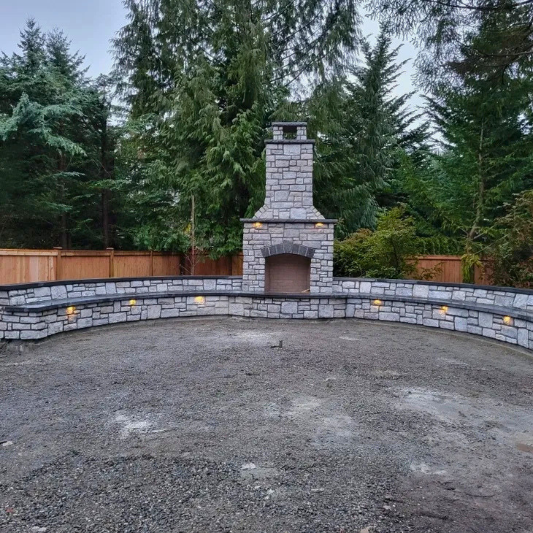 A backyard with a stone outdoor fireplace and a stone wall, surrounded by tall evergreen trees and a wooden fence in the background, on a cloudy day.