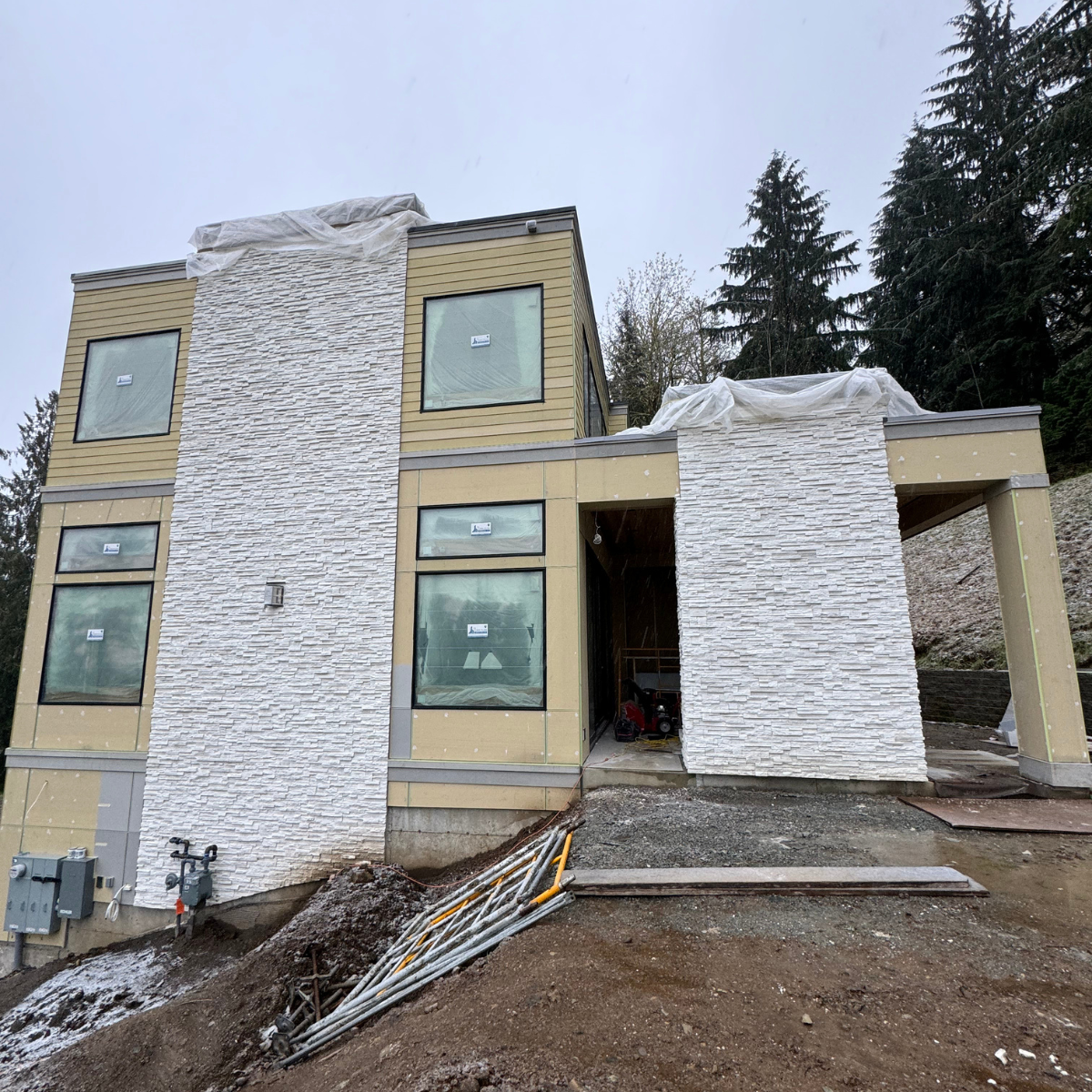 A modern house under construction with beige siding and white textured stone accents, large windows, and construction materials on the ground.