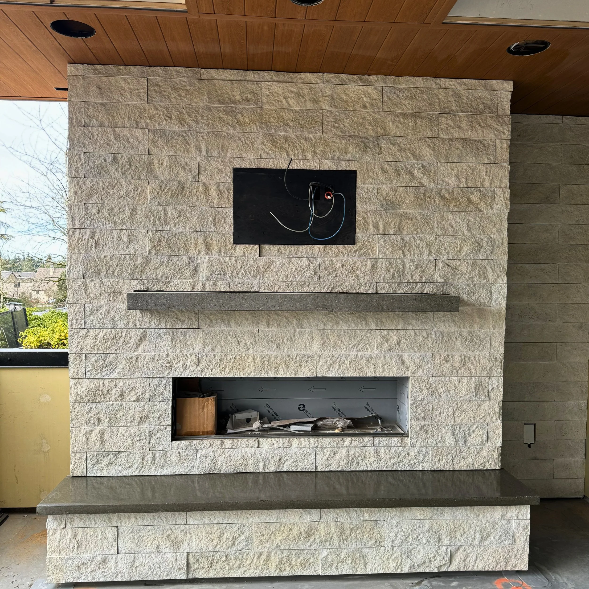 The image shows a partially built fireplace with exterior stone walls and an open space in the center for an insert. There are electrical wires in the opening, and construction materials are visible inside. The ceiling and part of the exterior wall are visible, indicating the fireplace is indoors.