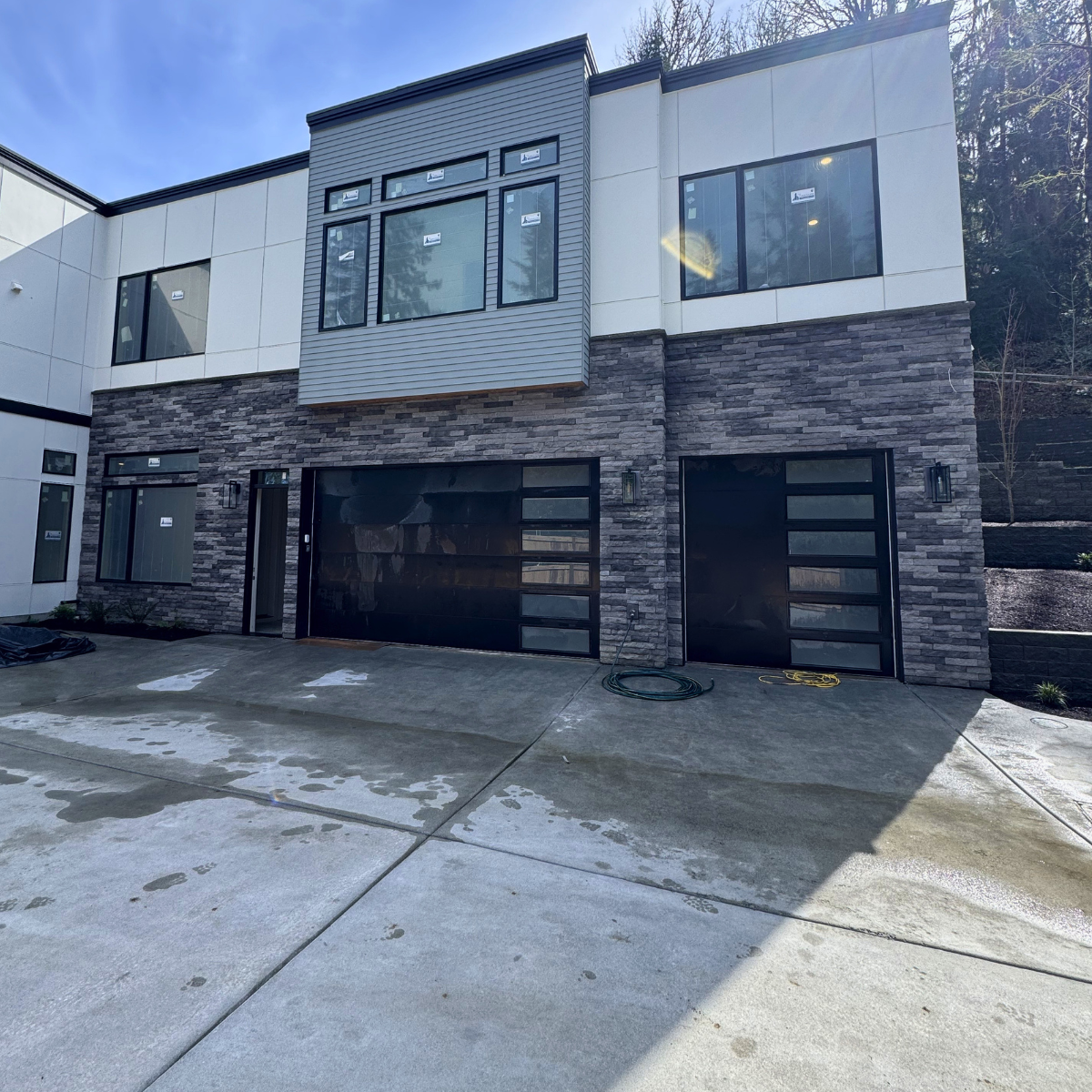 Newly built modern two-story house with a combination of stone and siding exterior, large windows, black garage doors, and a concrete driveway with some water spots.