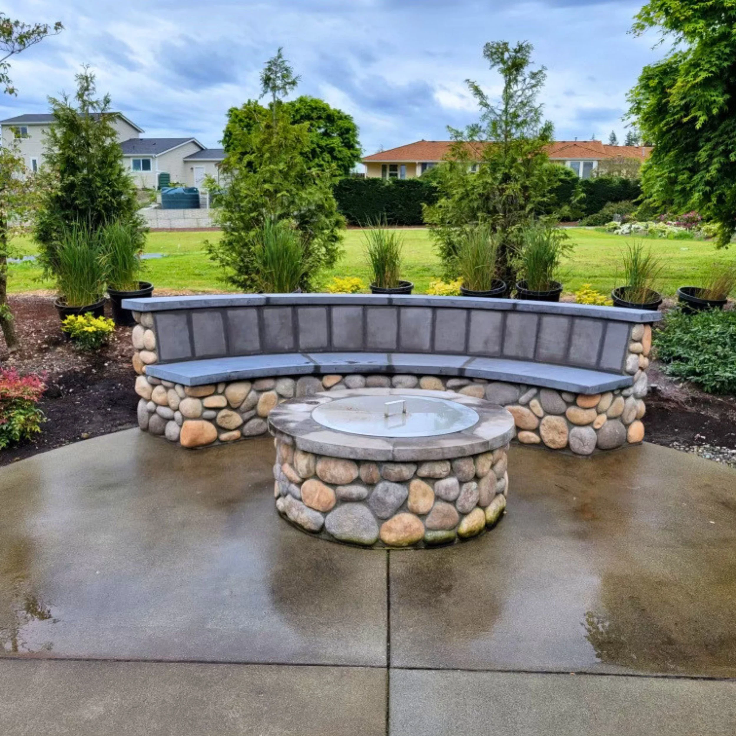 The image shows a curved stone and cement bench in the foreground, positioned on a concrete sidewalk. In the background, there are several young trees in pots and a well-maintained grassy area with shrubs. Beyond the greenery, there are several residential houses with white walls, te estás, and large windows. The sky is partly cloudy with some blue showing.