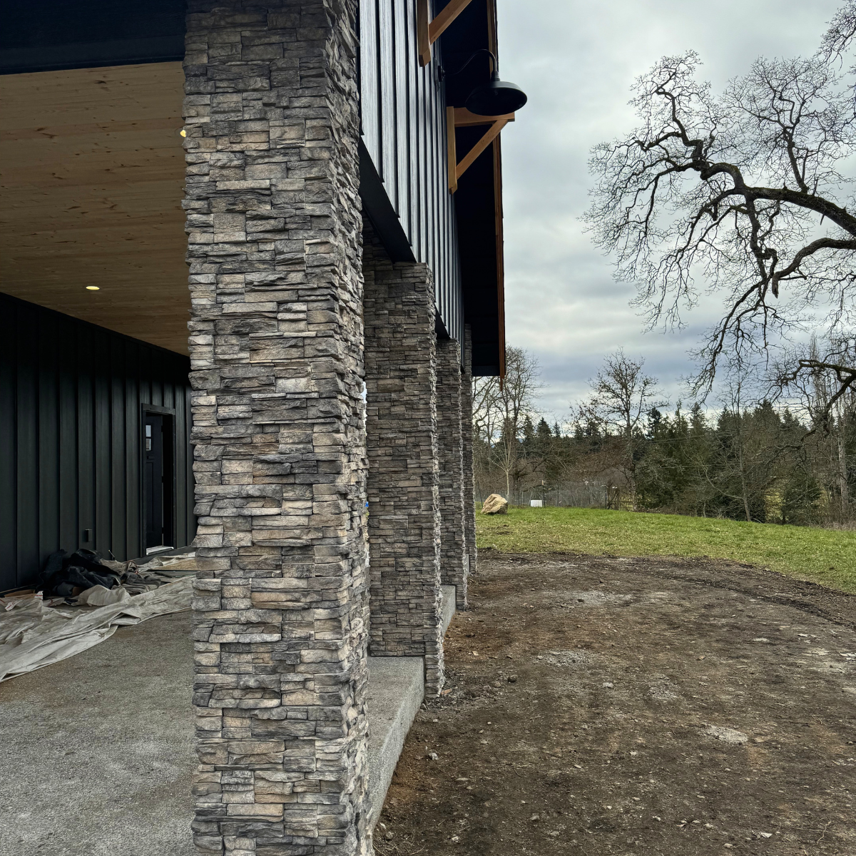 Partial view of a modern building with stone pillars supporting a wooden and metal overhang, overlooking a grassy, slightly muddy yard with leafless trees and a cloudy sky.