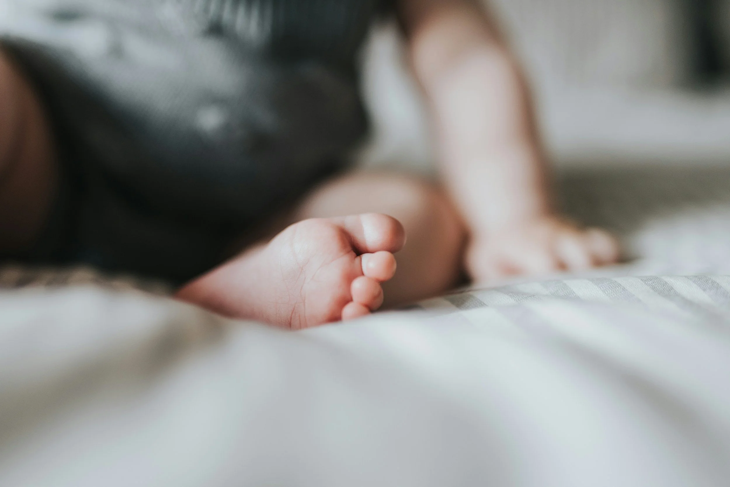 Close-up of a baby's foot lying on a bed with a blurred dark object in the background.