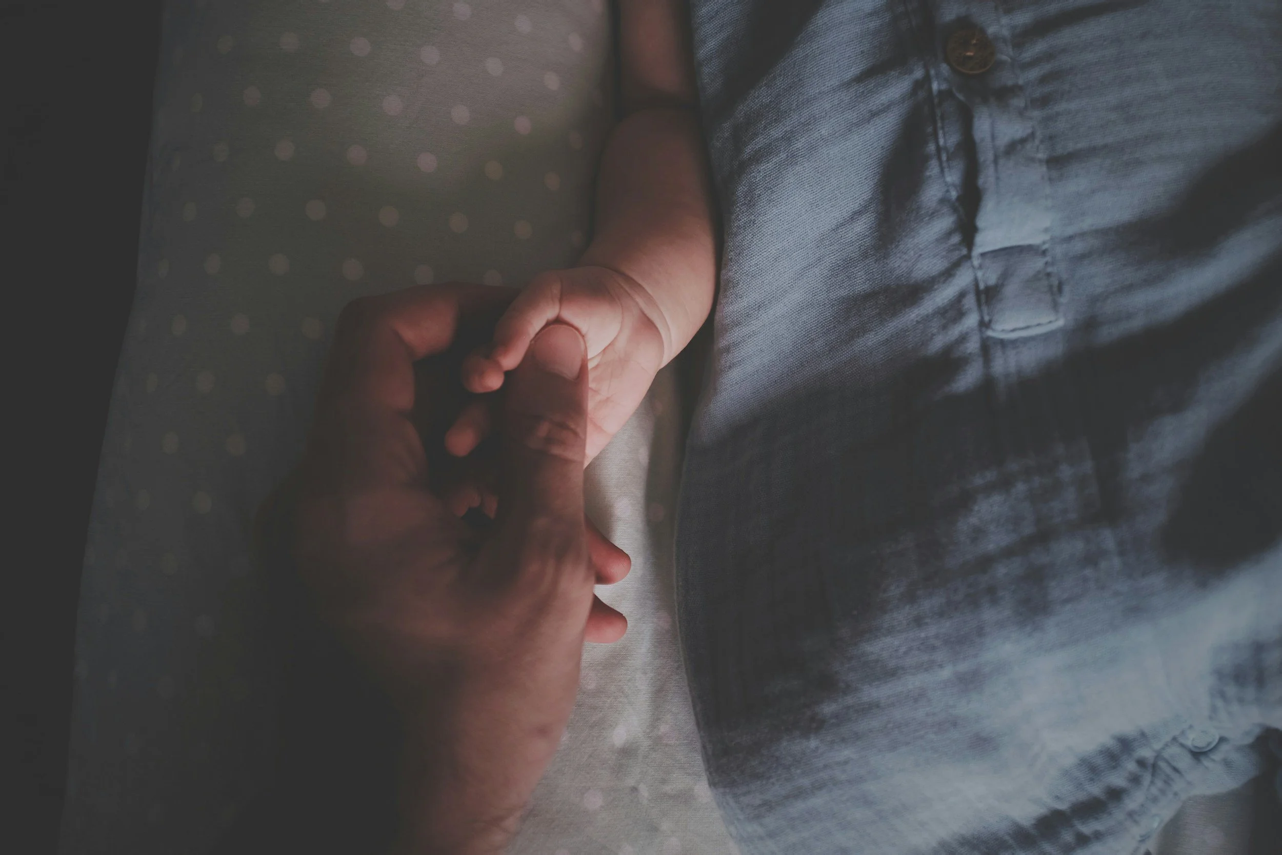 A close-up of a person's hand gently holding a baby's hand. The baby is dressed in a polka-dot onesie, and the adult is wearing a gray denim shirt.