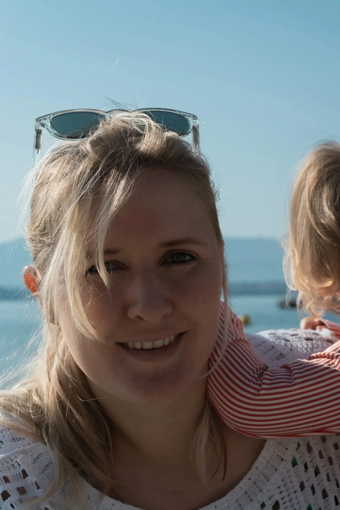 A woman with sunglasses on her head smiling at the camera, with a child on her shoulder near a beach or seaside.