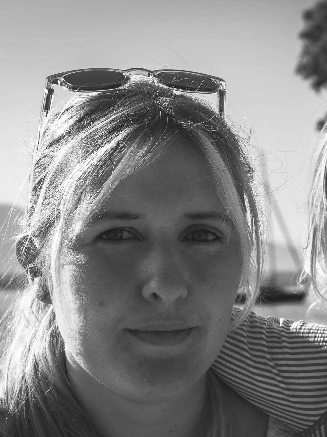 Close-up of a woman's face outdoors, wearing sunglasses on her head, with sunlight illuminating her hair and face, against a clear sky.