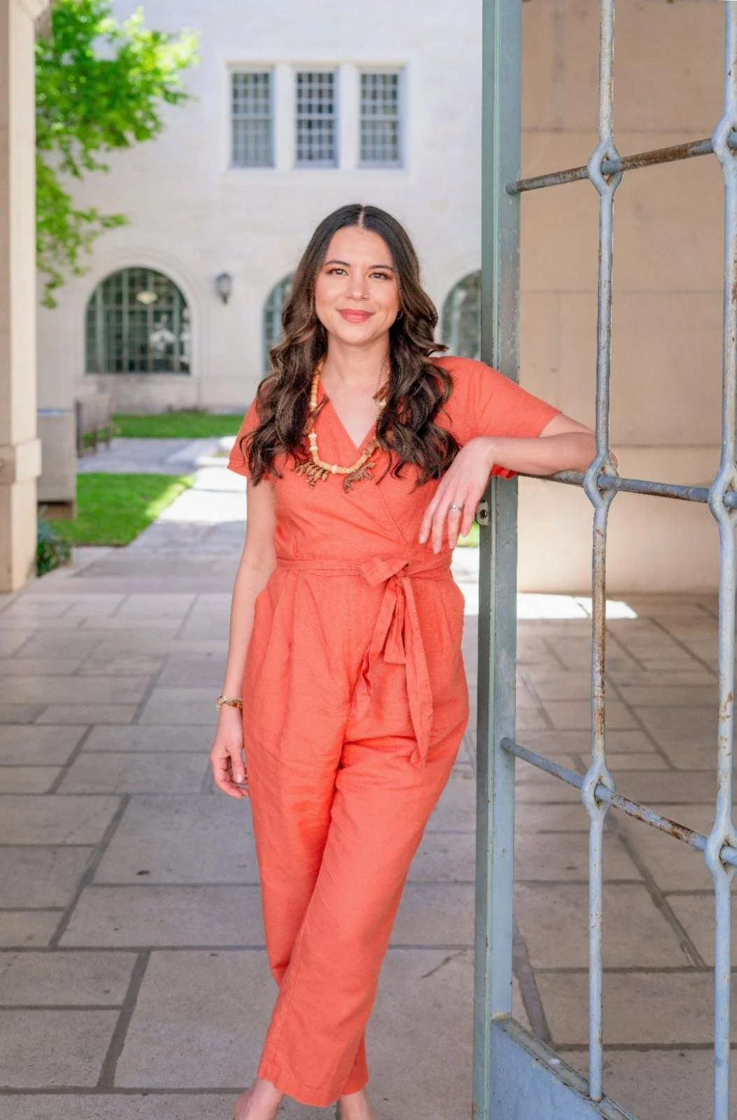A woman with long dark hair, wearing an orange jumpsuit and a beaded necklace, leaning on a metal gate outdoor on a sunny day.