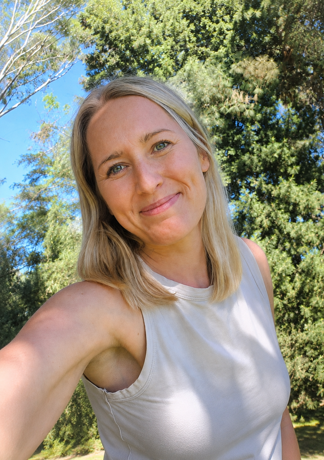 A woman with blonde hair and blue eyes smiling outdoors, wearing a light-colored sleeveless top with trees and blue sky in the background.