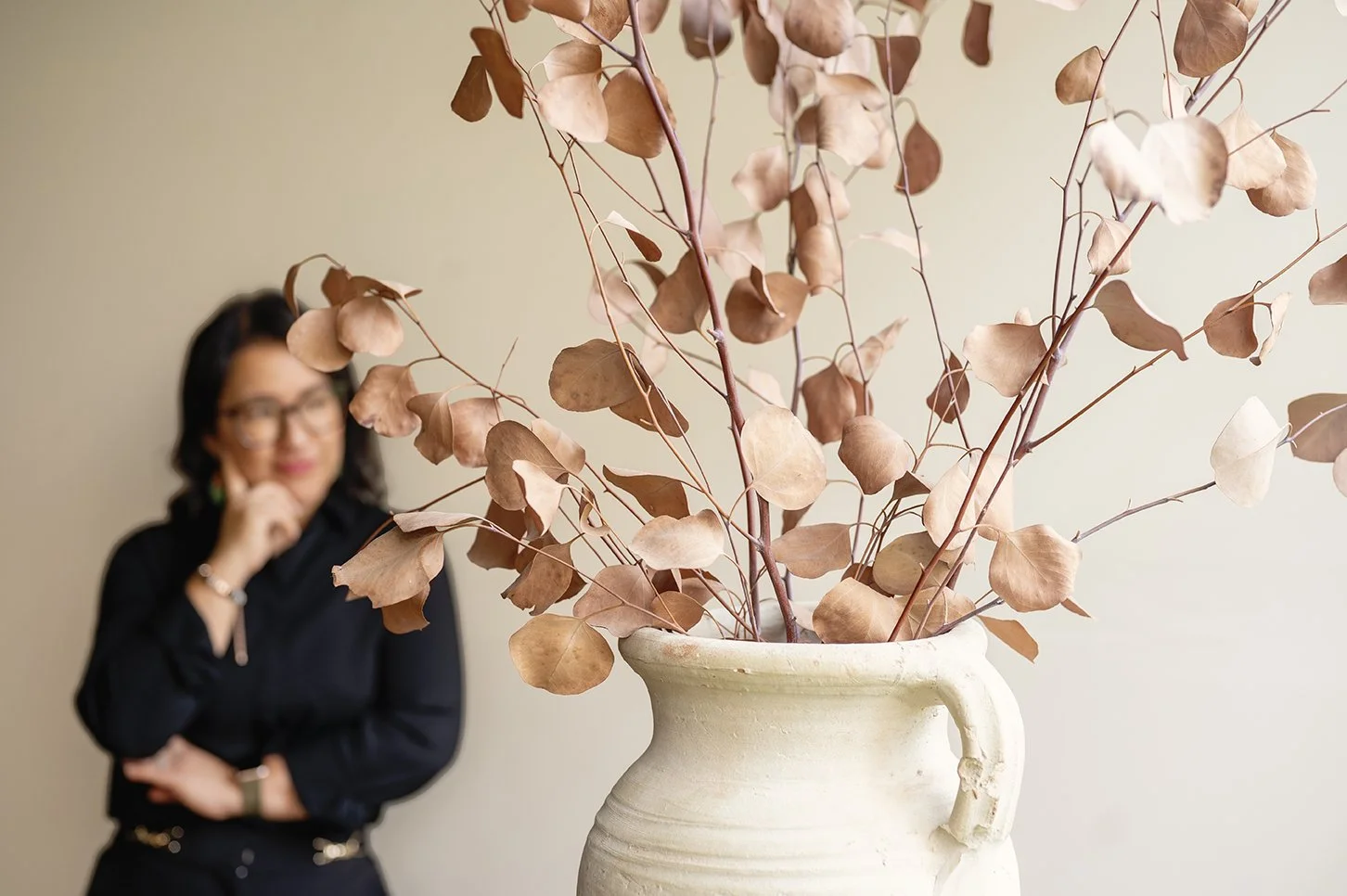 A large ceramic vase with dried pinkish-brown leaves and branches, with a woman in glasses and a black shirt blurred in the background.