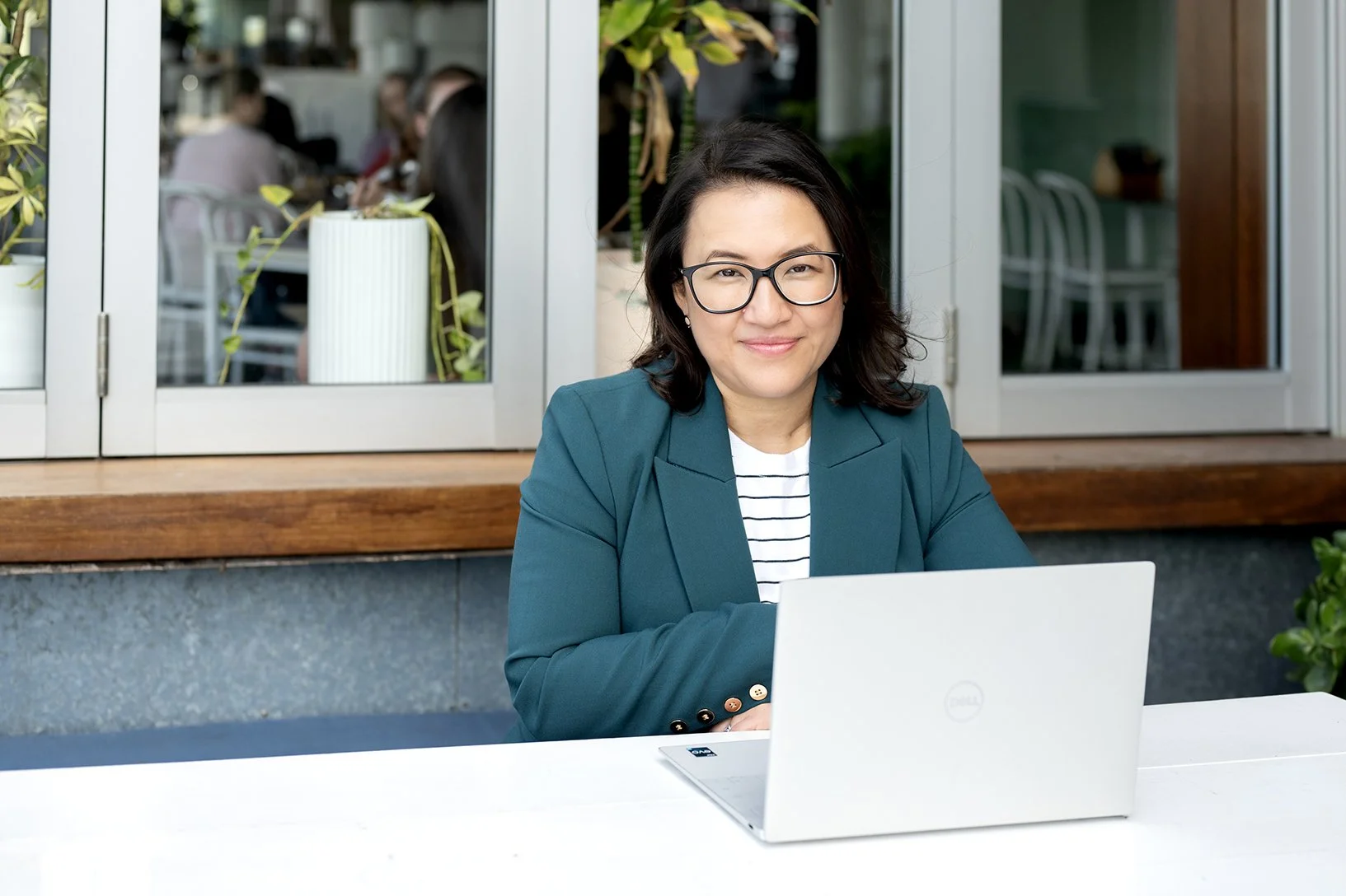 A woman with dark hair, glasses, and a teal blazer seated at a white table outside a cafe, smiling with a laptop in front of her.