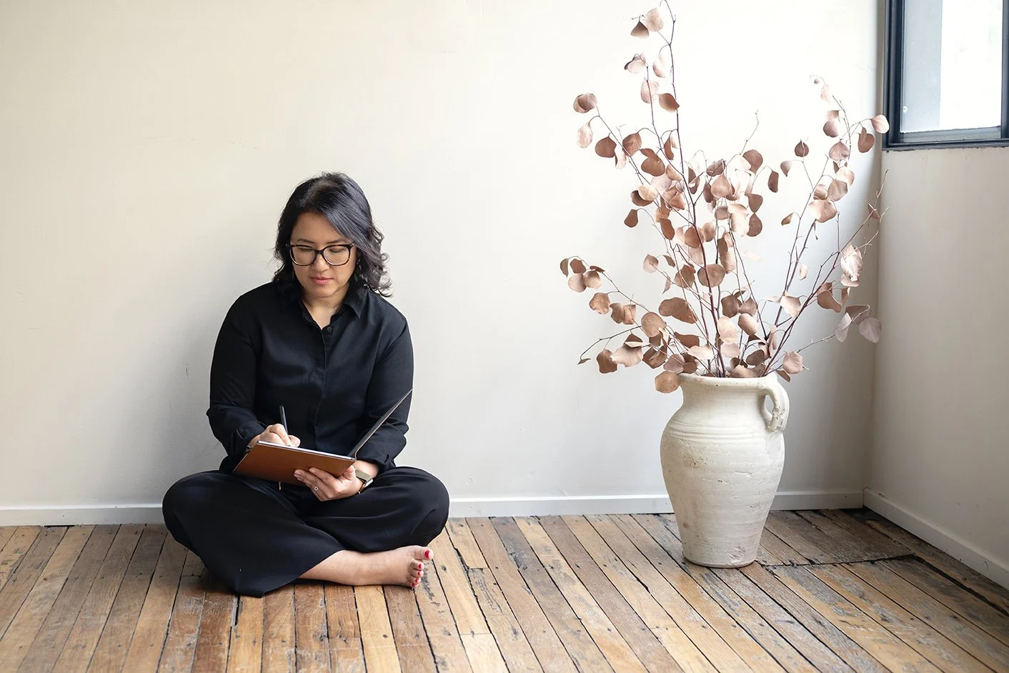 A woman with glasses and dark hair sitting cross-legged on a wooden floor, writing in a notebook. Next to her is a large cream-colored vase with dried pinkish-brown leaves. There is a window on the right side allowing natural light into the room.