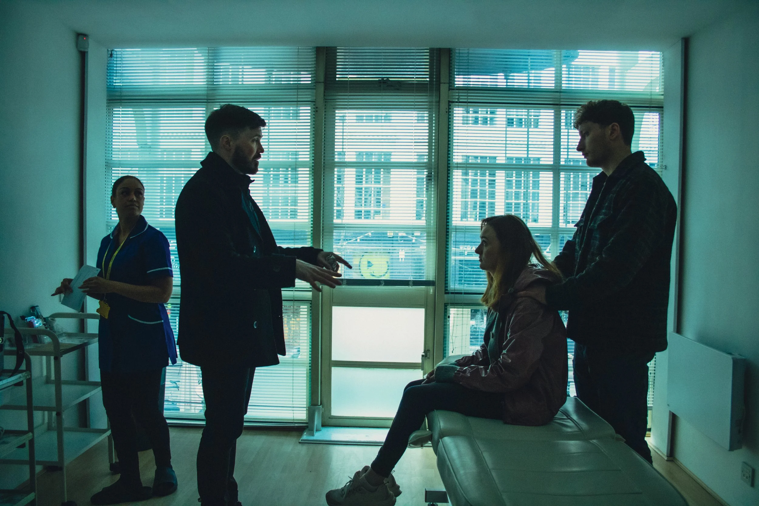 A hospital room with two young women sitting on hospital beds and a man and woman standing in front of them, talking. The room has large windows with blinds letting in natural light.