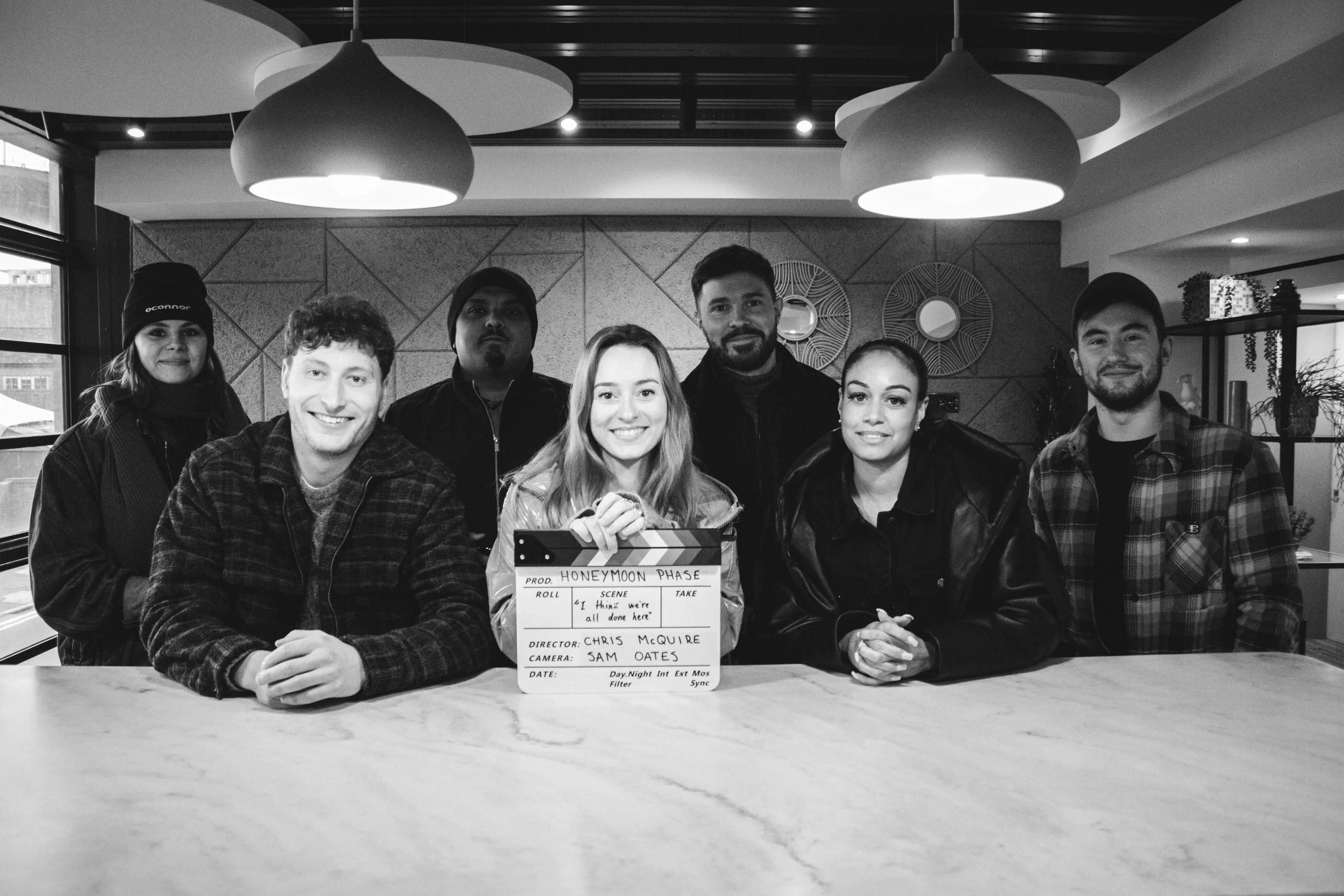 Group of seven young adults posing in a modern indoor setting with decorative wall art and large pendant lights. One person in the front holds a clapperboard labeled "HONEYMOON PHASE."