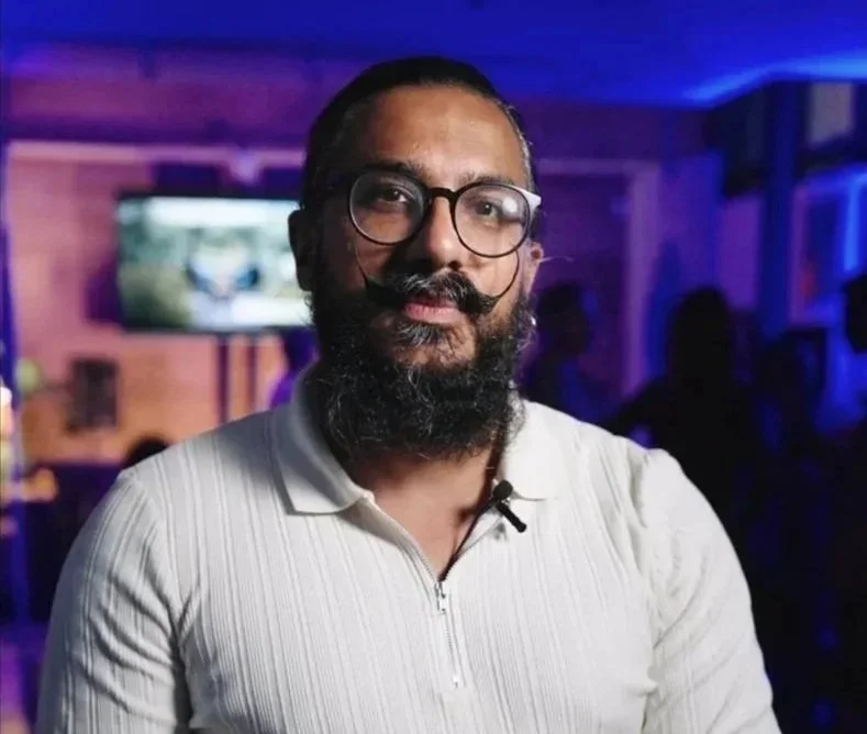 A man with glasses and a beard, wearing a white shirt, standing indoors with a blurred background of people and a television screen.