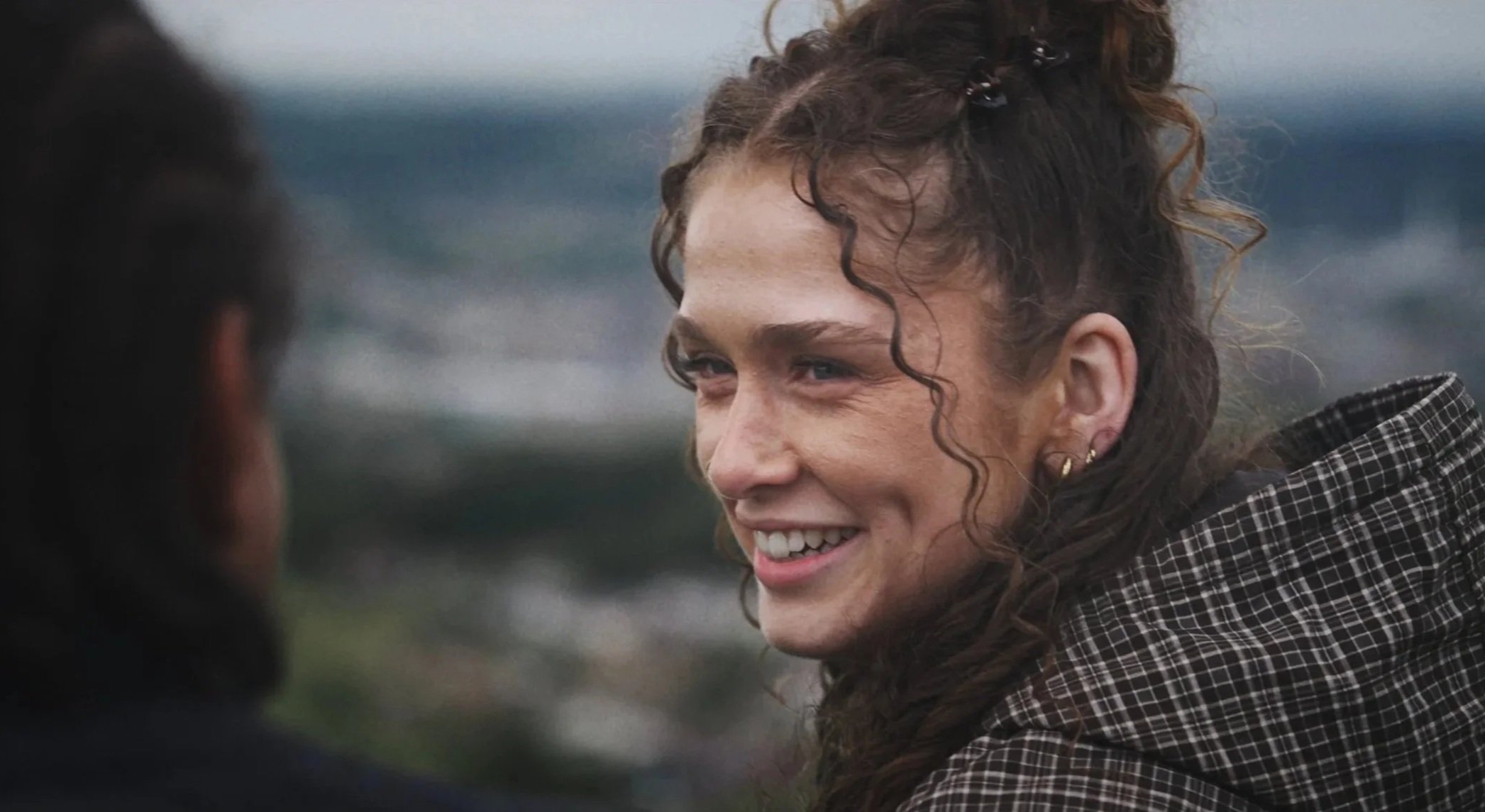A woman with curly hair and earrings smiling and looking at a man, with a blurry background.