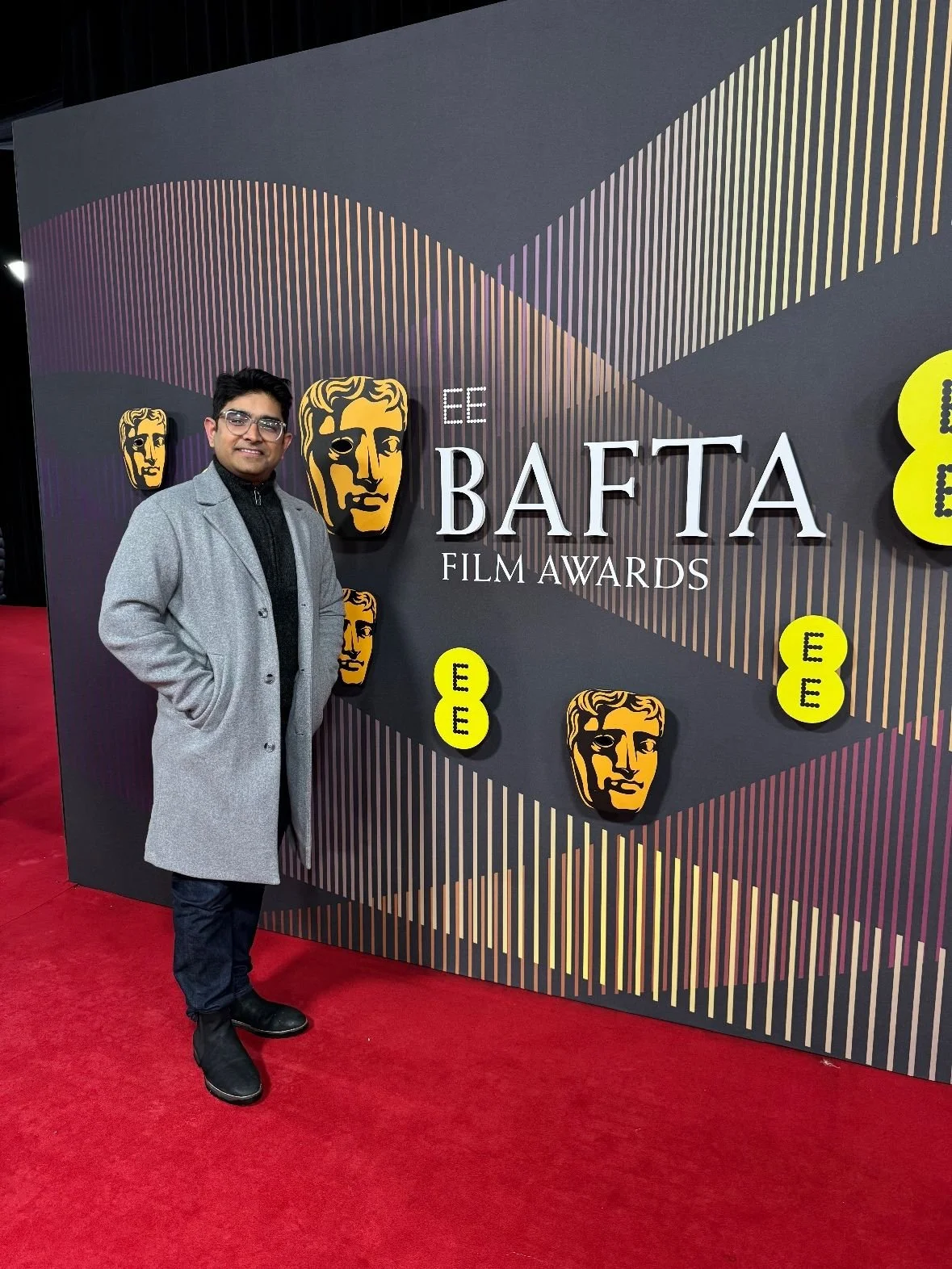 A man standing on a red carpet in front of a black backdrop with BAFTA film awards logos and symbols. The man is wearing glasses, a gray coat, black pants, and black shoes.