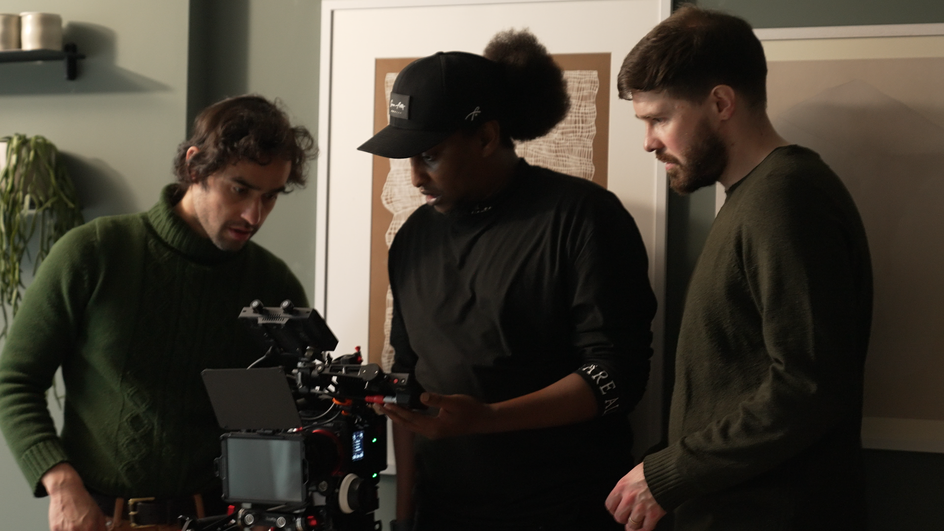 Three people examining a movie camera in a room, with a green wall and a plant in the background.