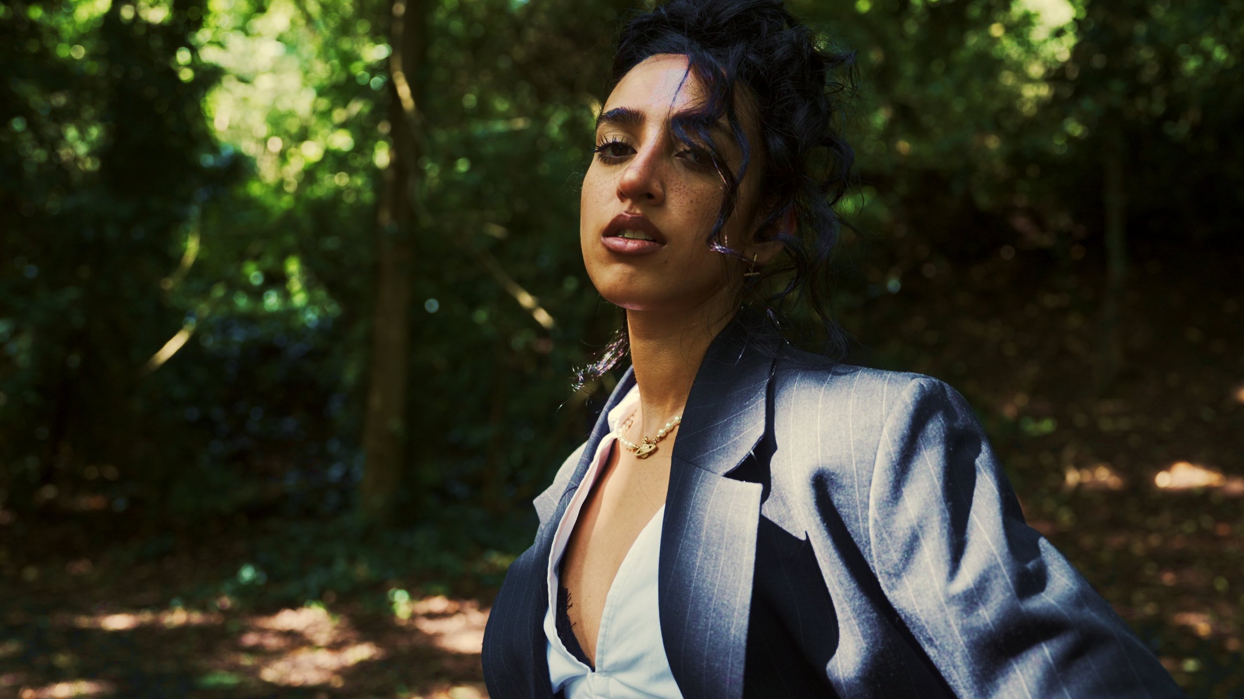 A woman with dark, curly hair and freckles wearing a pinstripe blazer, a pearl necklace, and a white shirt in a wooded area.