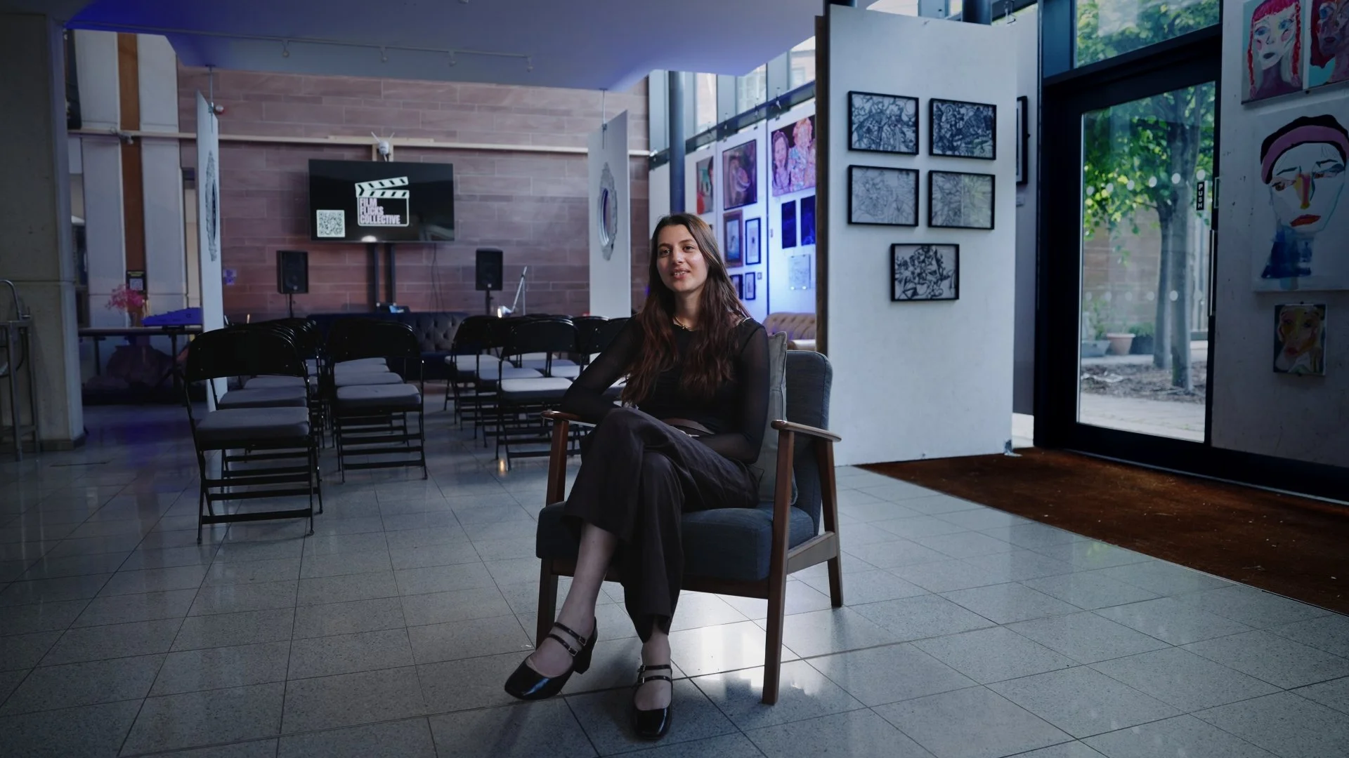 Interior of an art gallery with a woman sitting on a chair near the entrance, surrounded by framed artwork on the walls and a screen displaying a film clapperboard logo in the background, with chairs arranged for an event.